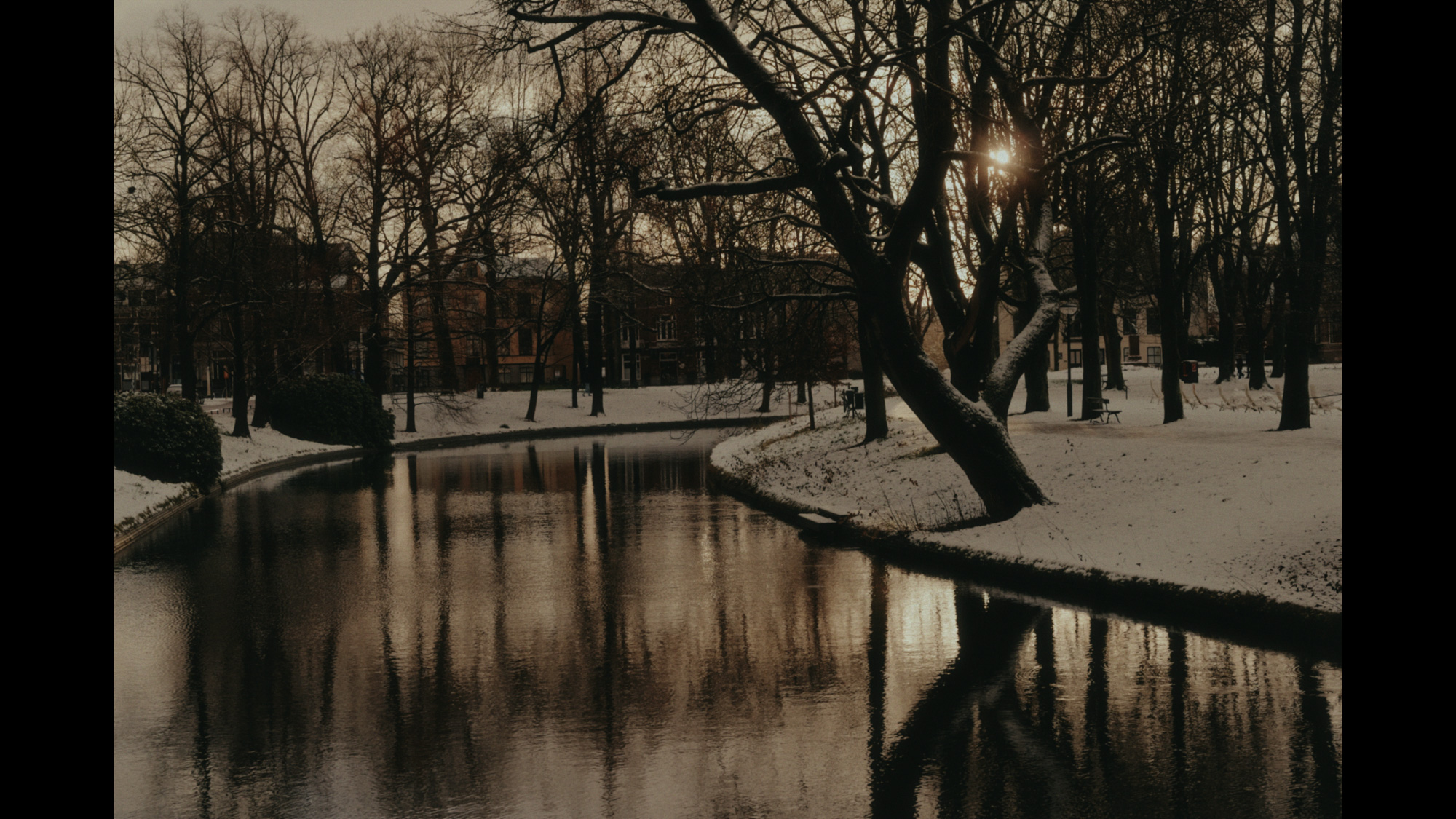 A snow-covered park features a winding canal reflecting bare trees and distant buildings under a muted winter sky.