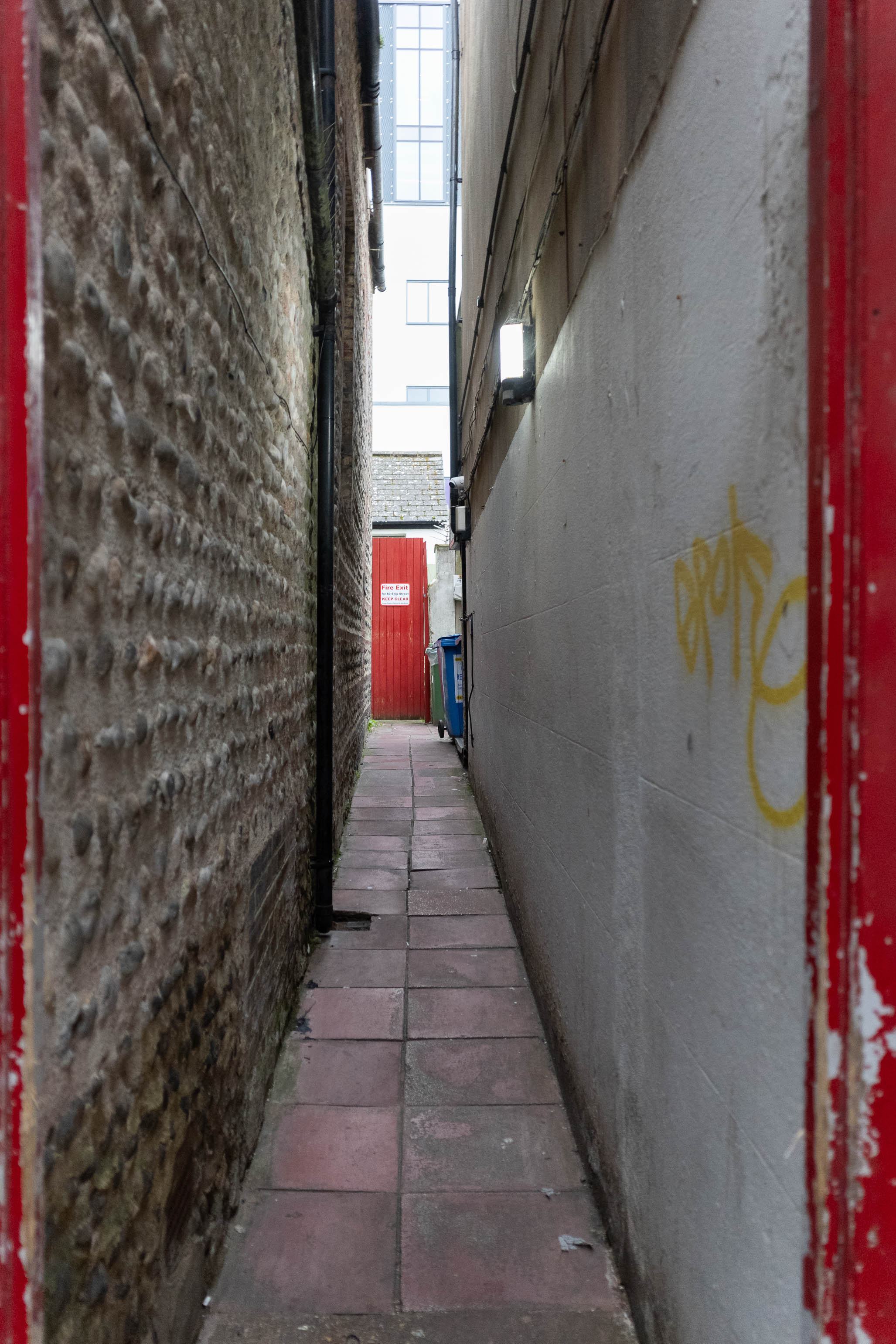 Dunkle Gasse mit roten Bodenplatten, die zu einer roten Holztür führen. Aufgenommen durch einen roten Türrahmen mit einer Ricoh GR IV.