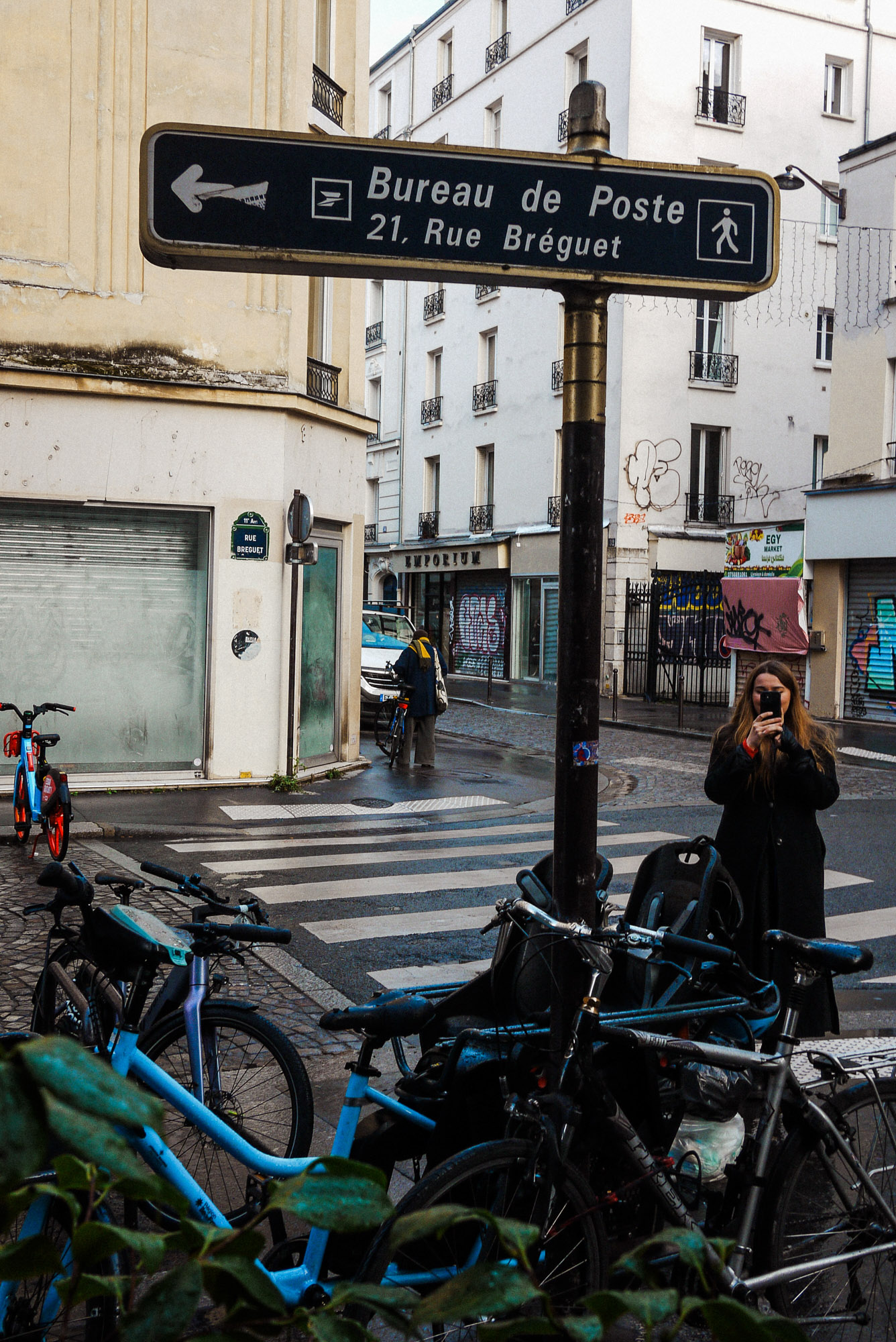 Streetaufnahme an einer Pariser Straßenecke mit einem Wegweiserschild zum Bureau de Poste in der Rue Bréguet, geparkten Fahrrädern und einer Person, die im Hintergrund ein Foto macht. Aufgenommen von Wesley Verhoeve mit einer Panasonic Lumix GF1.