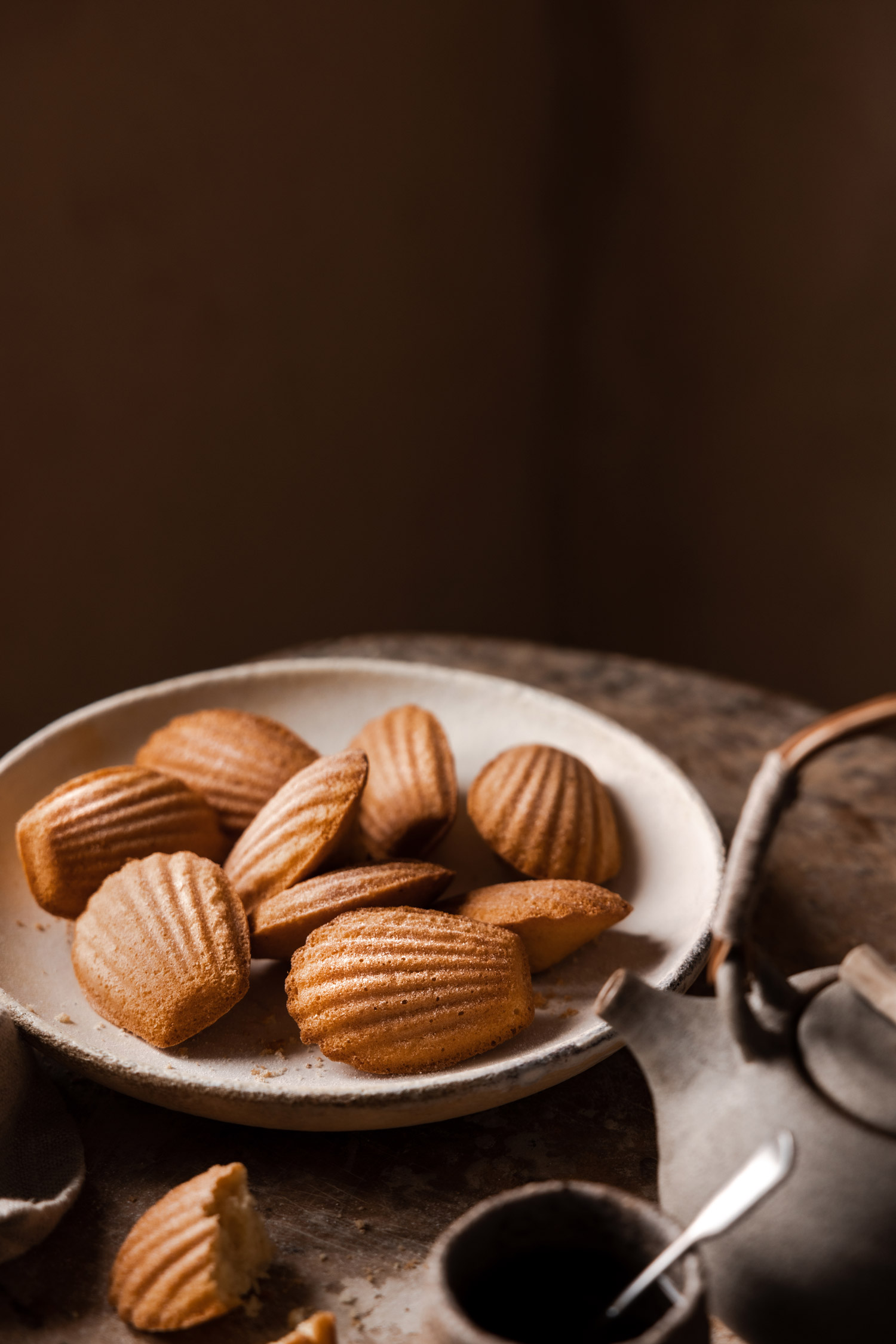 Madeleines sur une assiette blanche avec une théière et un sucrier au premier plan, prises par Bea Lubas avec le Canon EOS R5.

