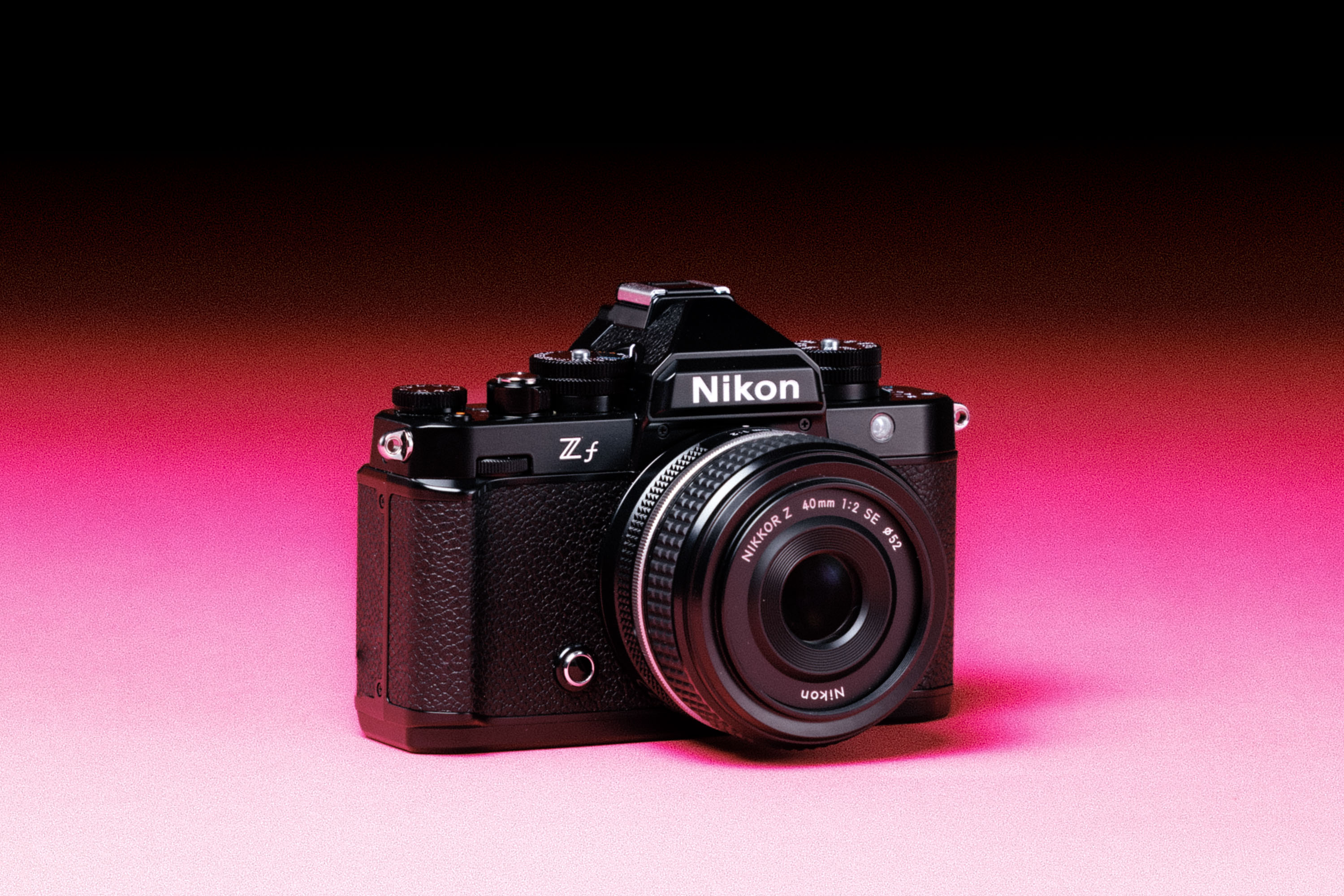 A used Nikon Z f with a Z 40mm f/2 lens, with an added film-like grain effect on a graduated pink background.