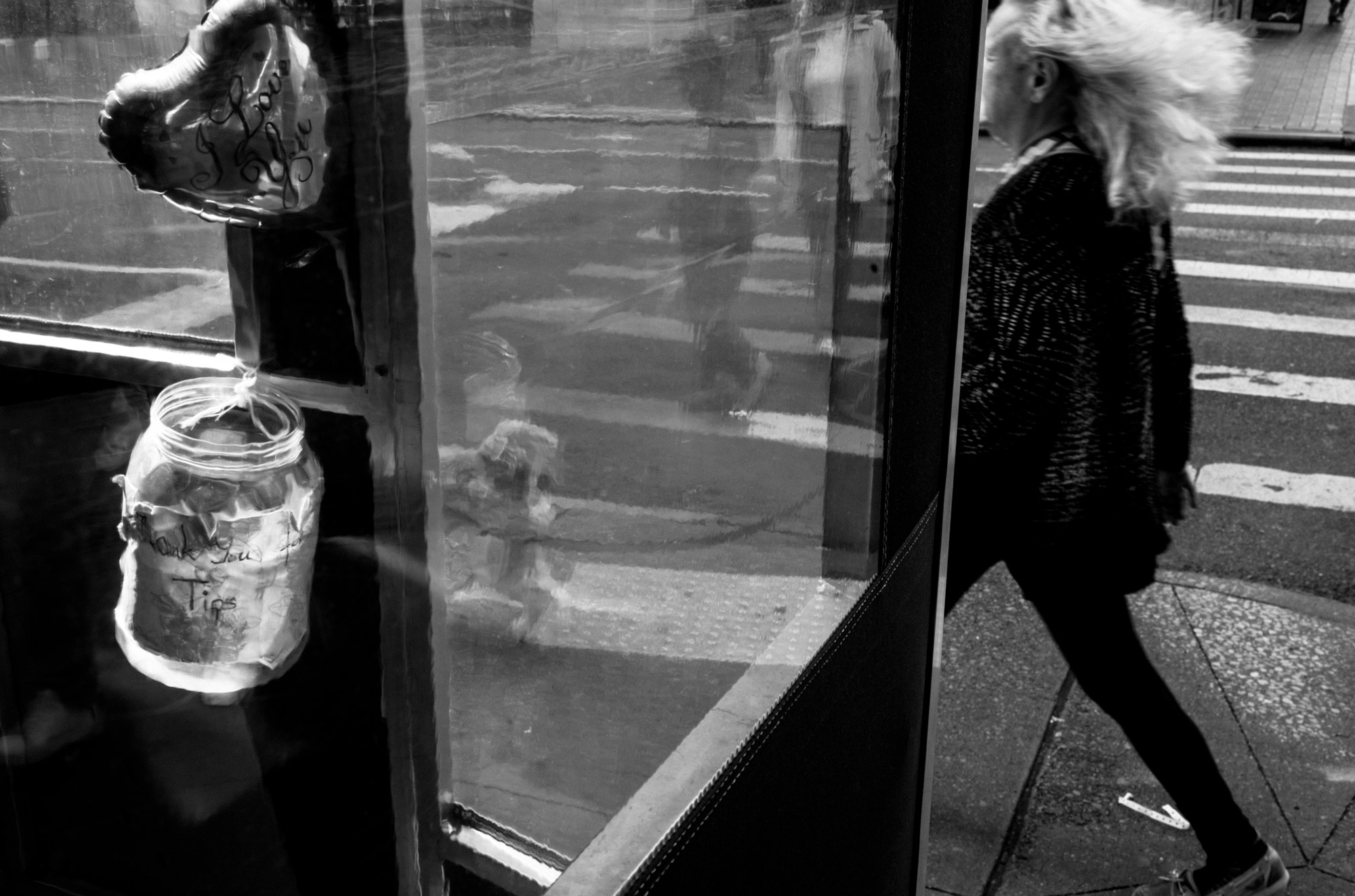 A black-and-white photo of a person walking their dog by a building with a heart balloon and a tip jar