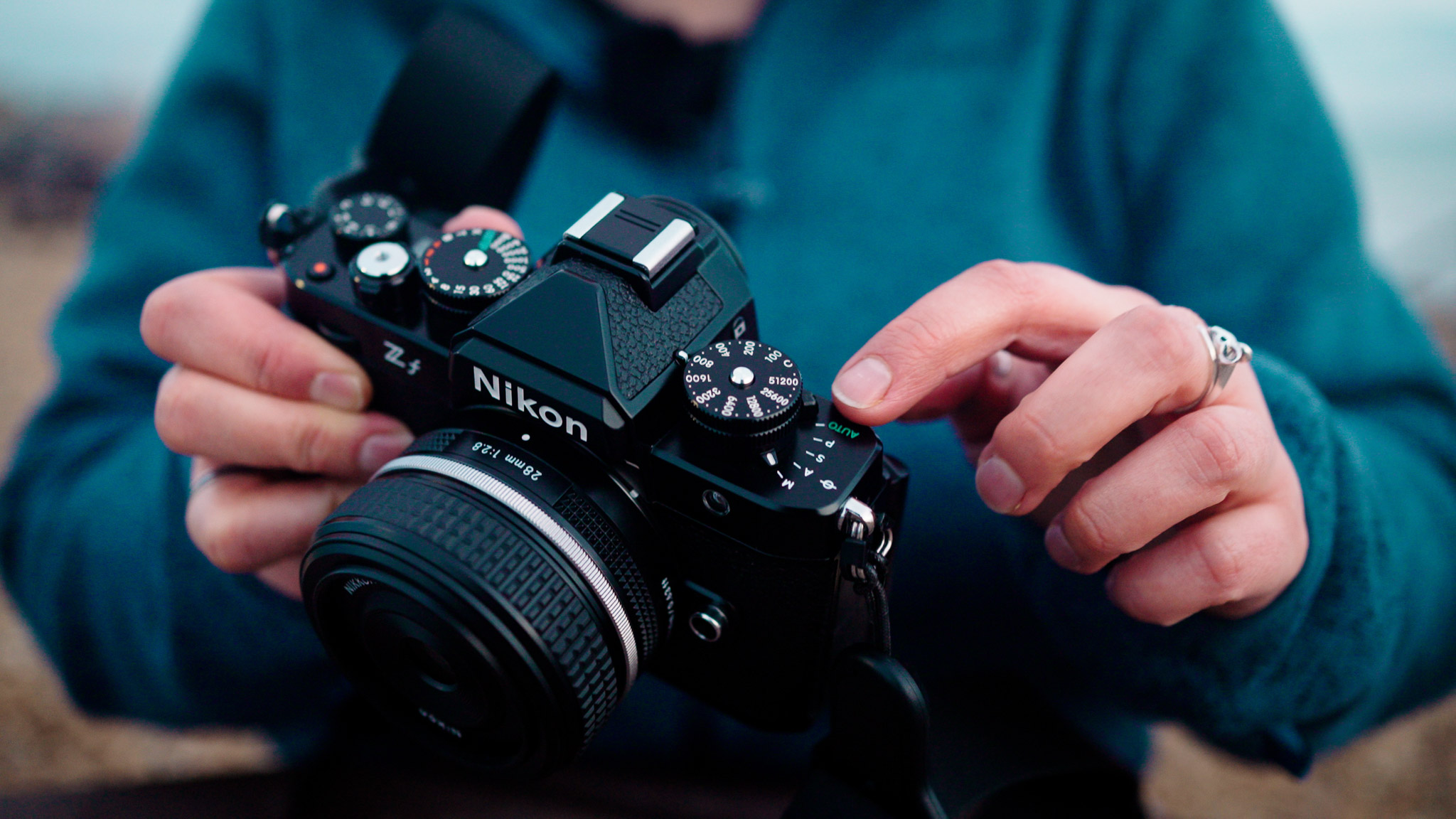 Close-up of a Nikon Zf full-frame mirrorless camera being held, showing the vintage-style top dials and mounted 28mm lens.