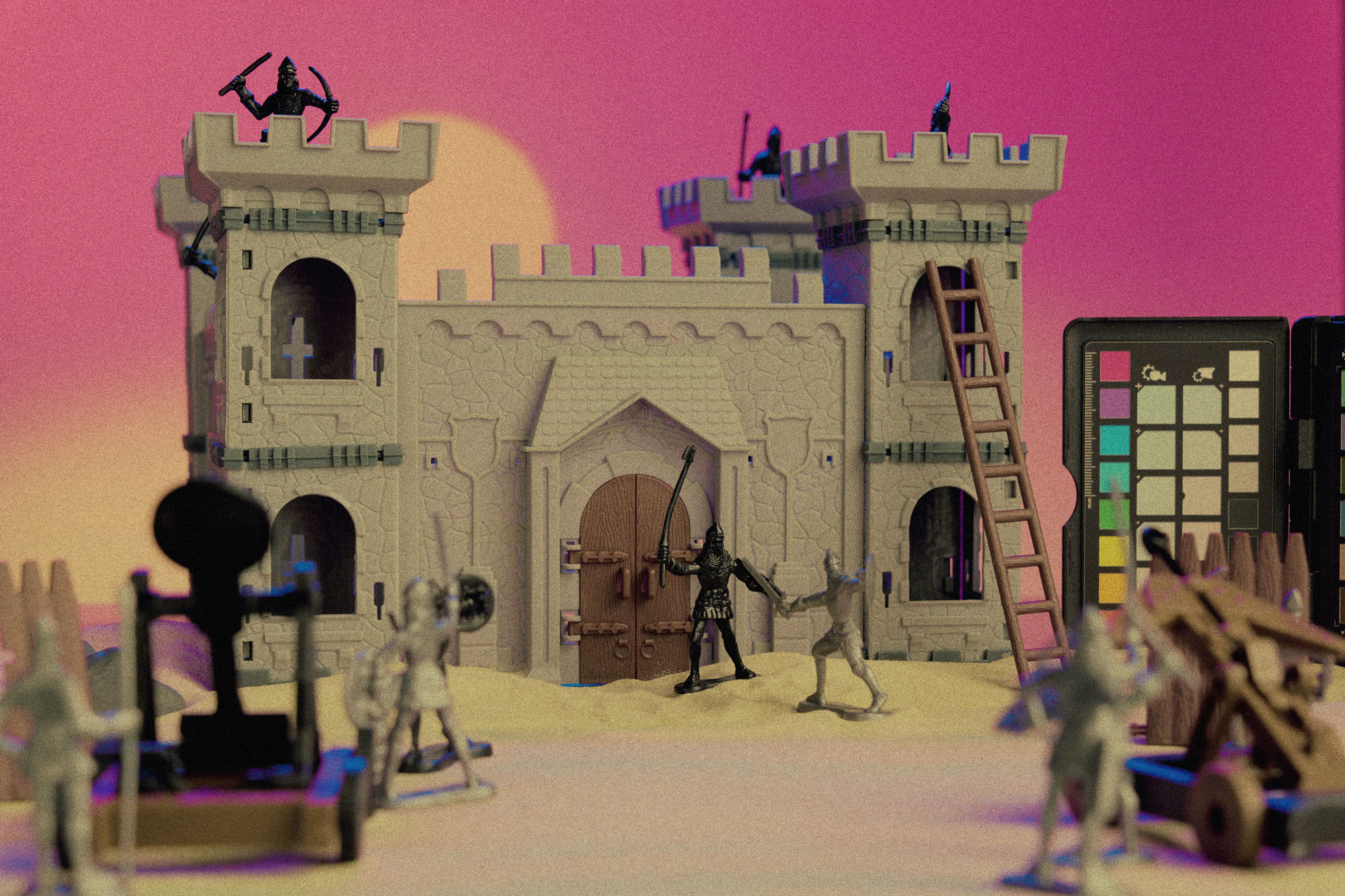 An example of a sample LUT. A model castle with knight figurines and a model ladder propped up on the side. There is a soft pink backdrop with soft light in the background. A Calibrite Colorchecker is coming in from the right.