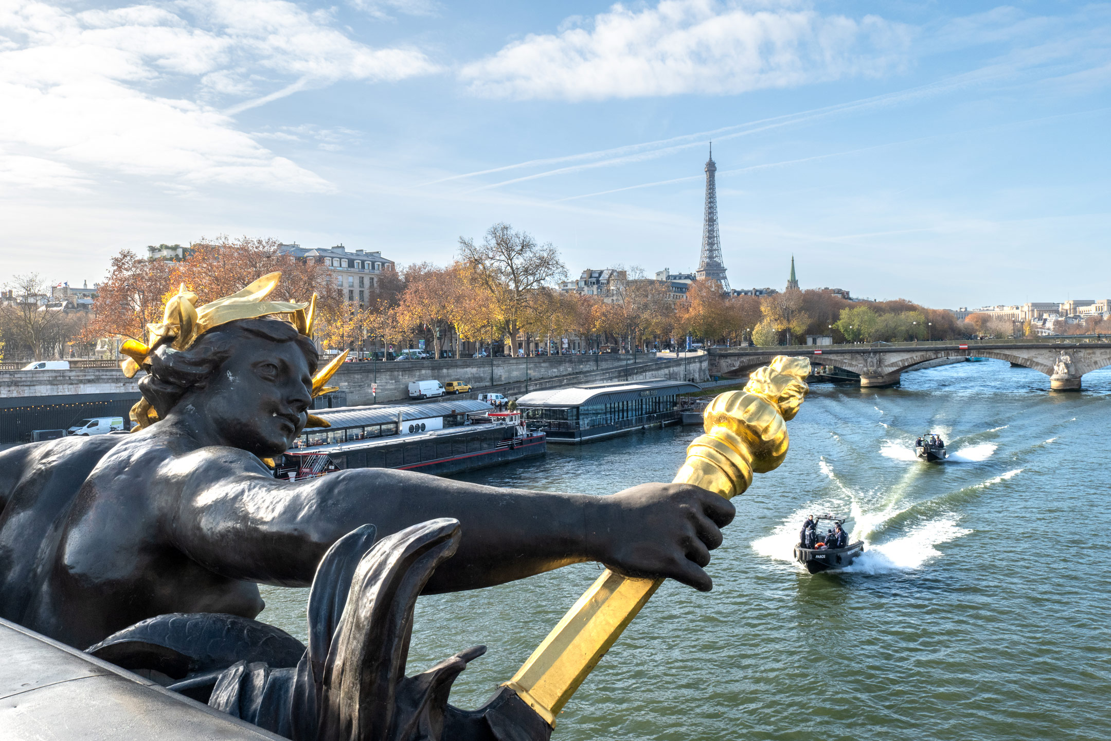 Golden statue on Pont Alexandre III overlooking the River Seine with boats and the Eiffel Tower in Paris, framed by autumn trees under a bright blue sky.