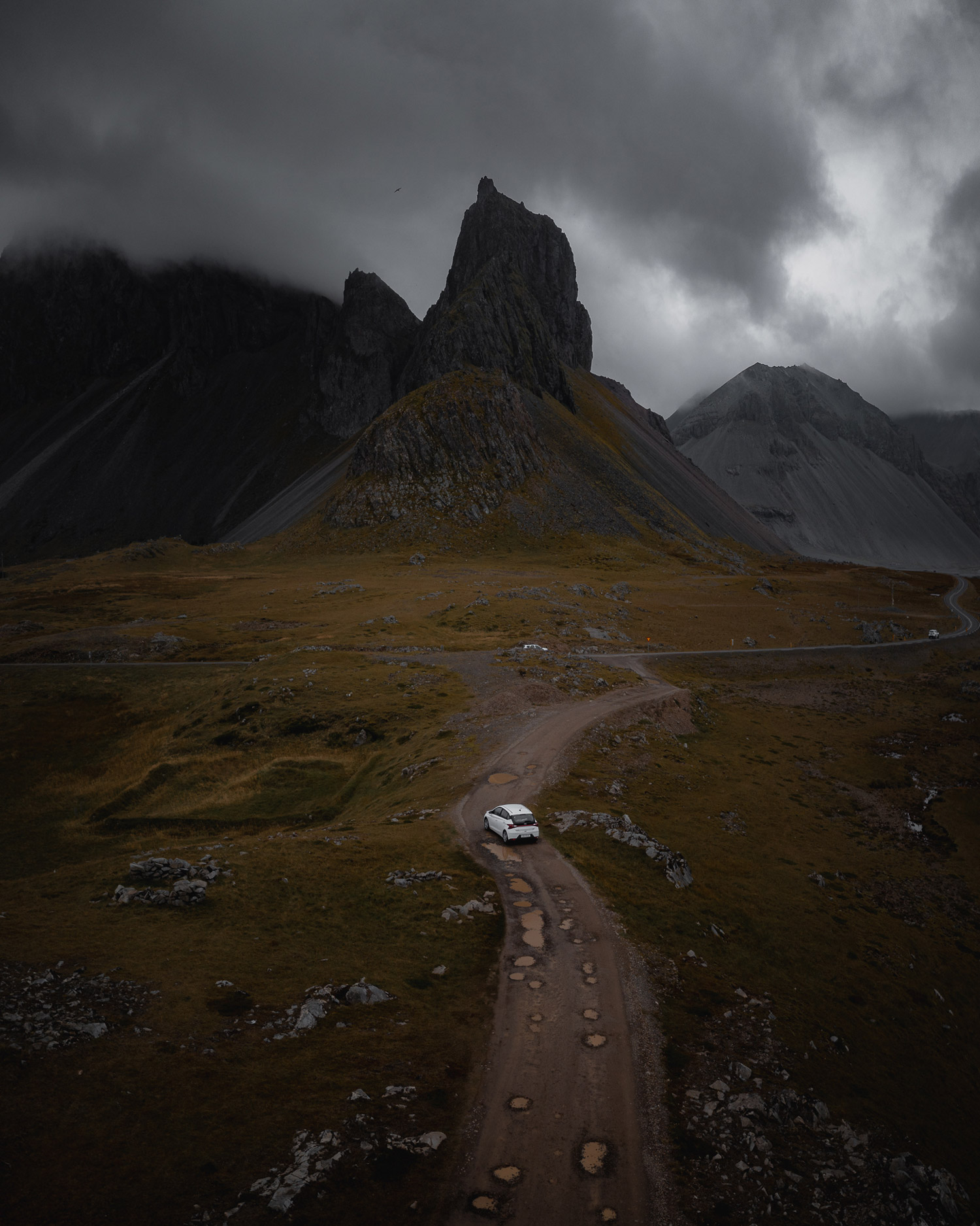 Aerial drone photo of a white car driving on a gravel road in Iceland, with mountains in the background and fields on either side of the road, shot by Paul Watson with a DJI Mavic 2 Pro