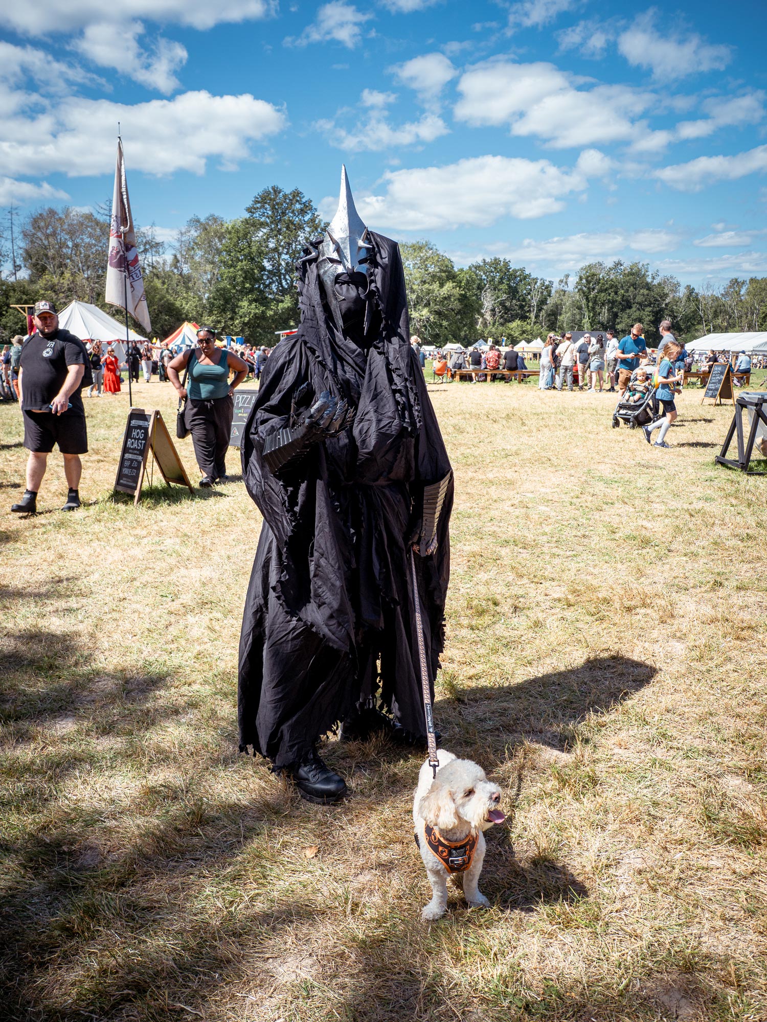 A person dressed as a fictional Nazgul stands at a medieval fair with his cockapoo on a lead. It is a sunny day.