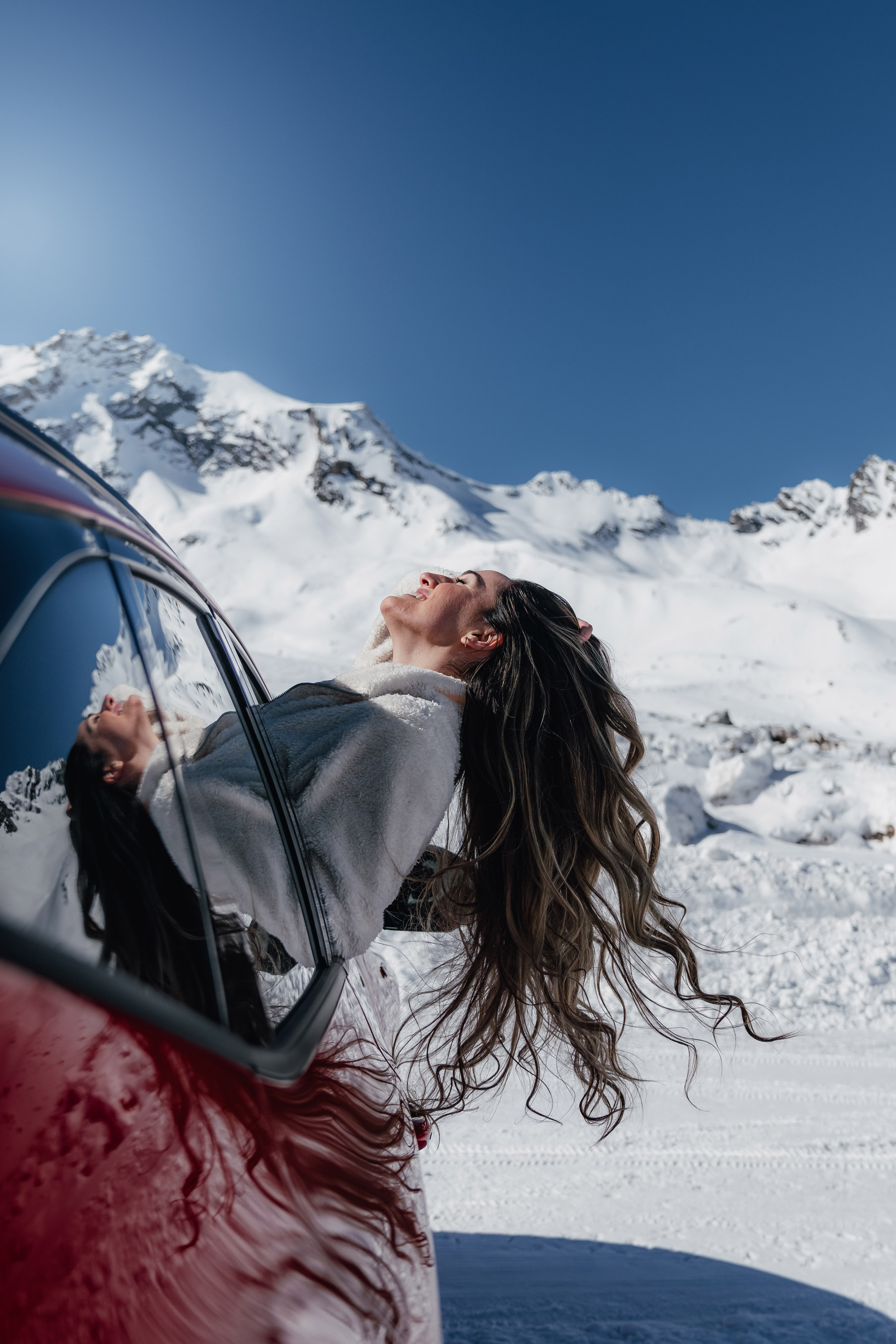 Woman leans head out of red car surrounded by snowy mountain on top of a glacier in Austria
