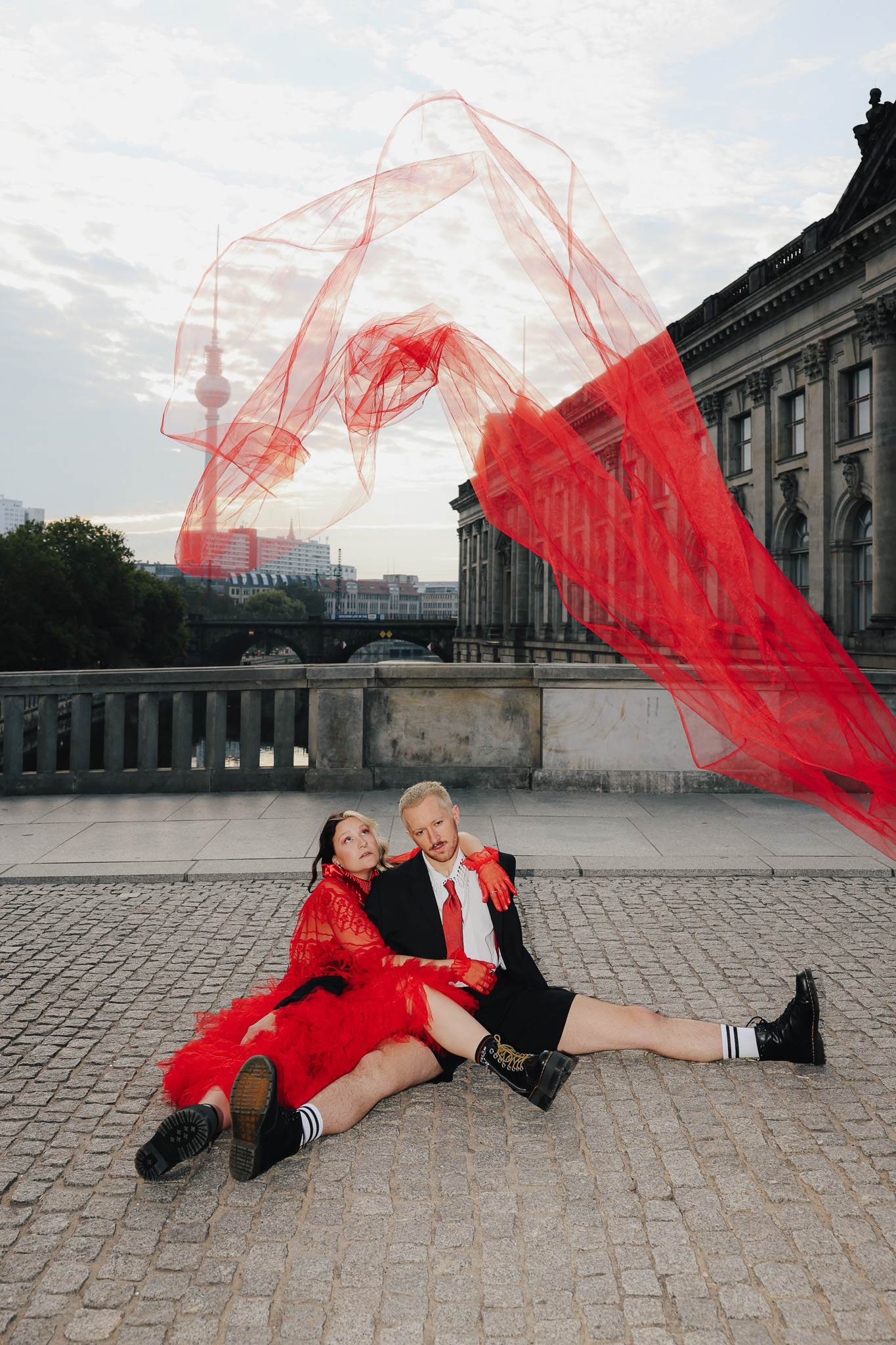 A creative wedding picture showing a couple sitting on the pavement, with Berlin landmarks in the background and a dramatic red veil in the foreground. Shot by Marina Polovinkina on a Canon EOS R6 Mark II.