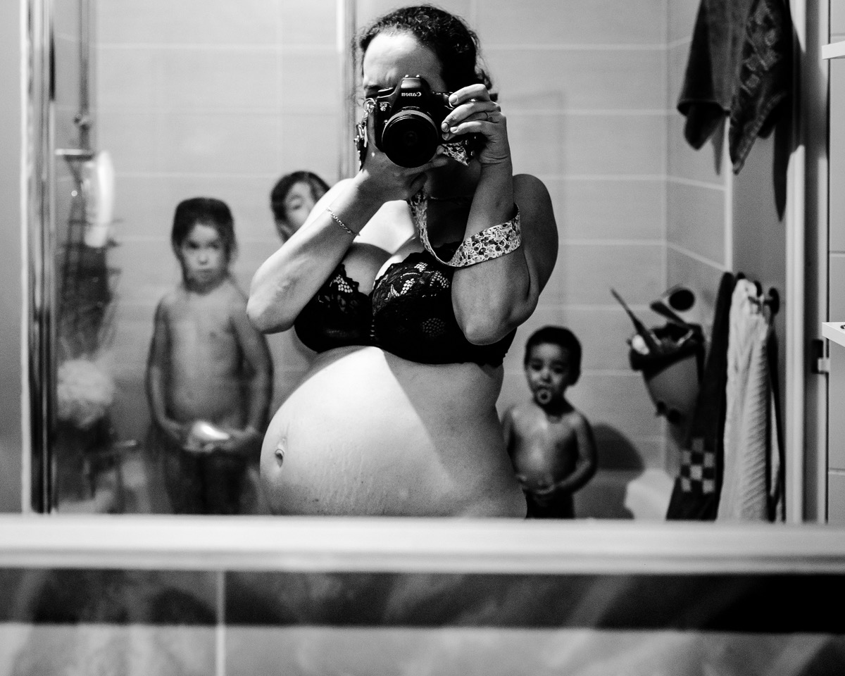 Une photo en noir et blanc d'une personne enceinte se prenant en photo dans le miroir de sa salle de bain, alors que ses enfants sont dans la baignoire derrière elle, prise par Rocio Marin Perez avec un Canon EOS 6D.