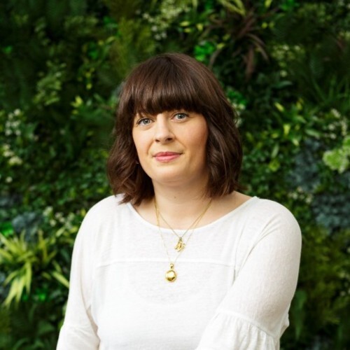 A photo of a lady with brown hair sitting in front of green garden wall.