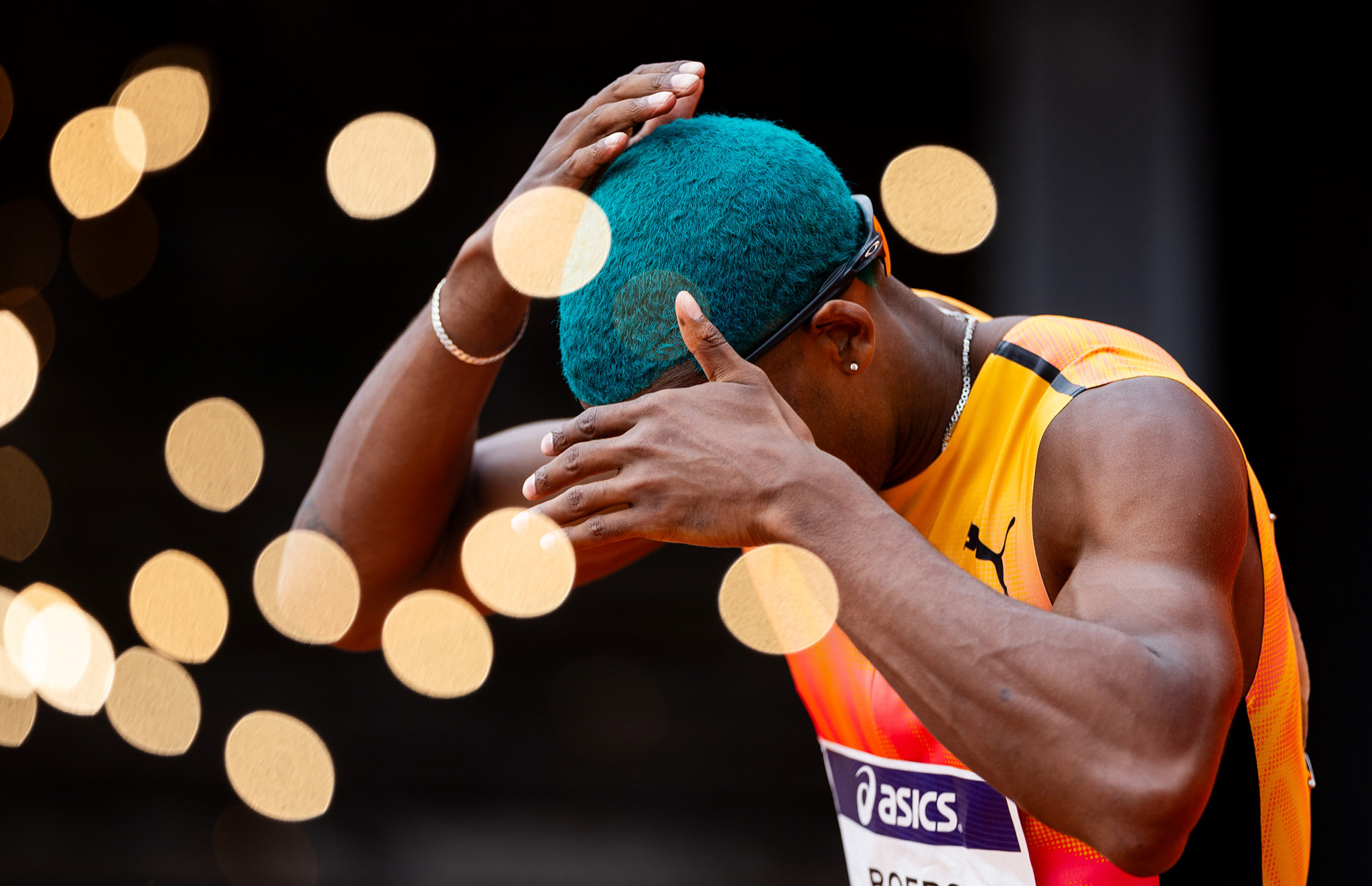 An athlete brushing their blue hair with their hands with bright yellow bokeh balls around them. By Iris van den Broek.