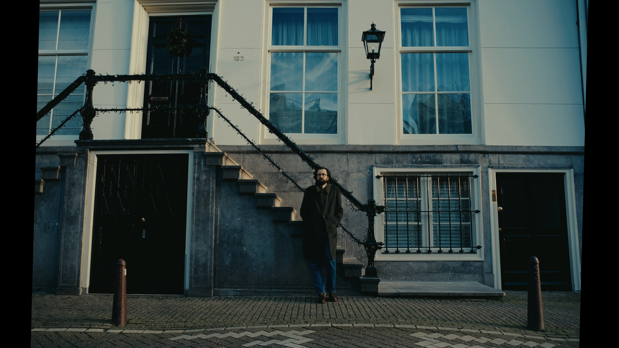 A person in a dark coat and glasses stands on the stone steps of an Amsterdam townhouse.
