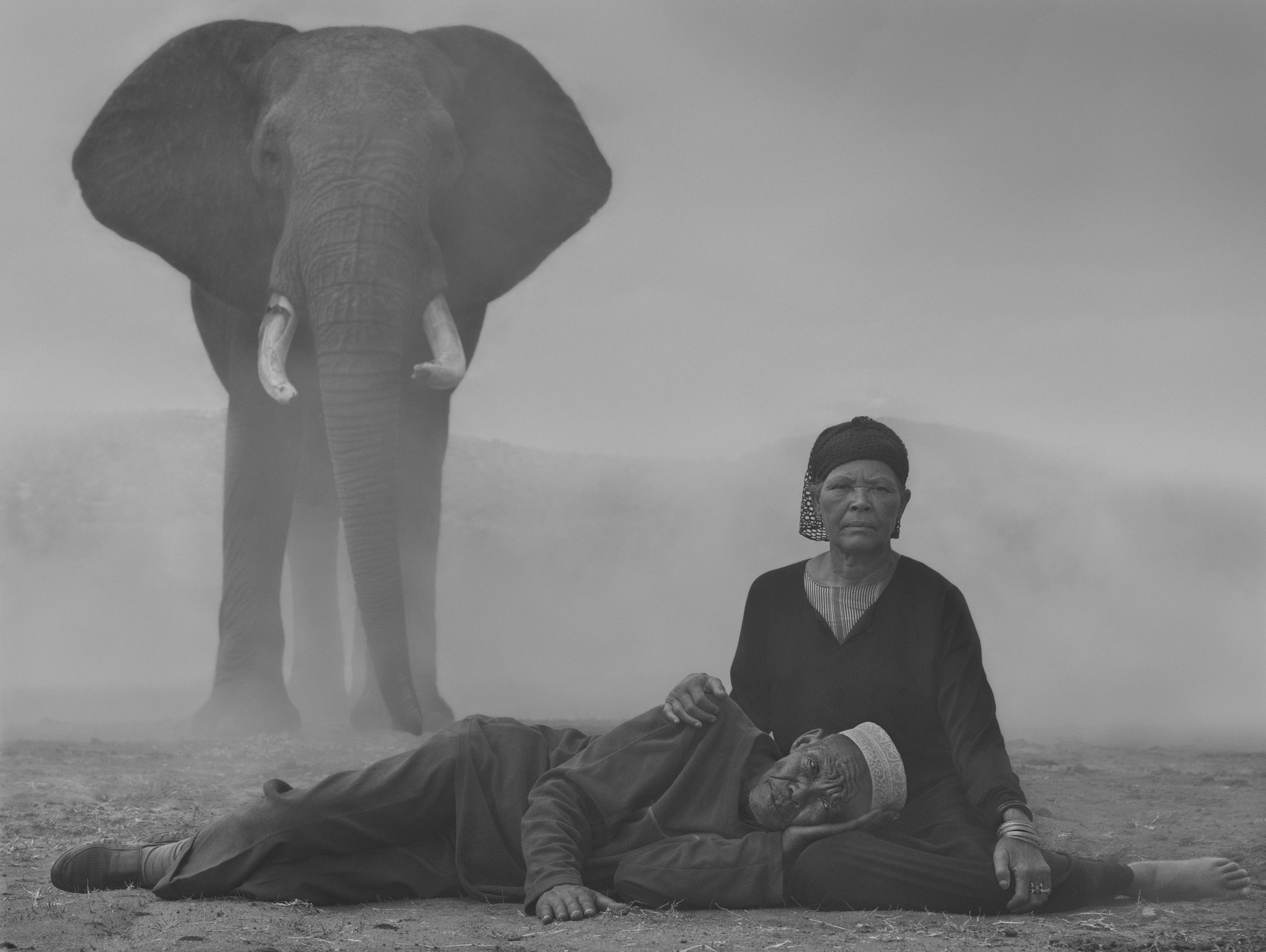 A black-and-white person lying down on the lap of another person in the desert, with an elephant in the background 
