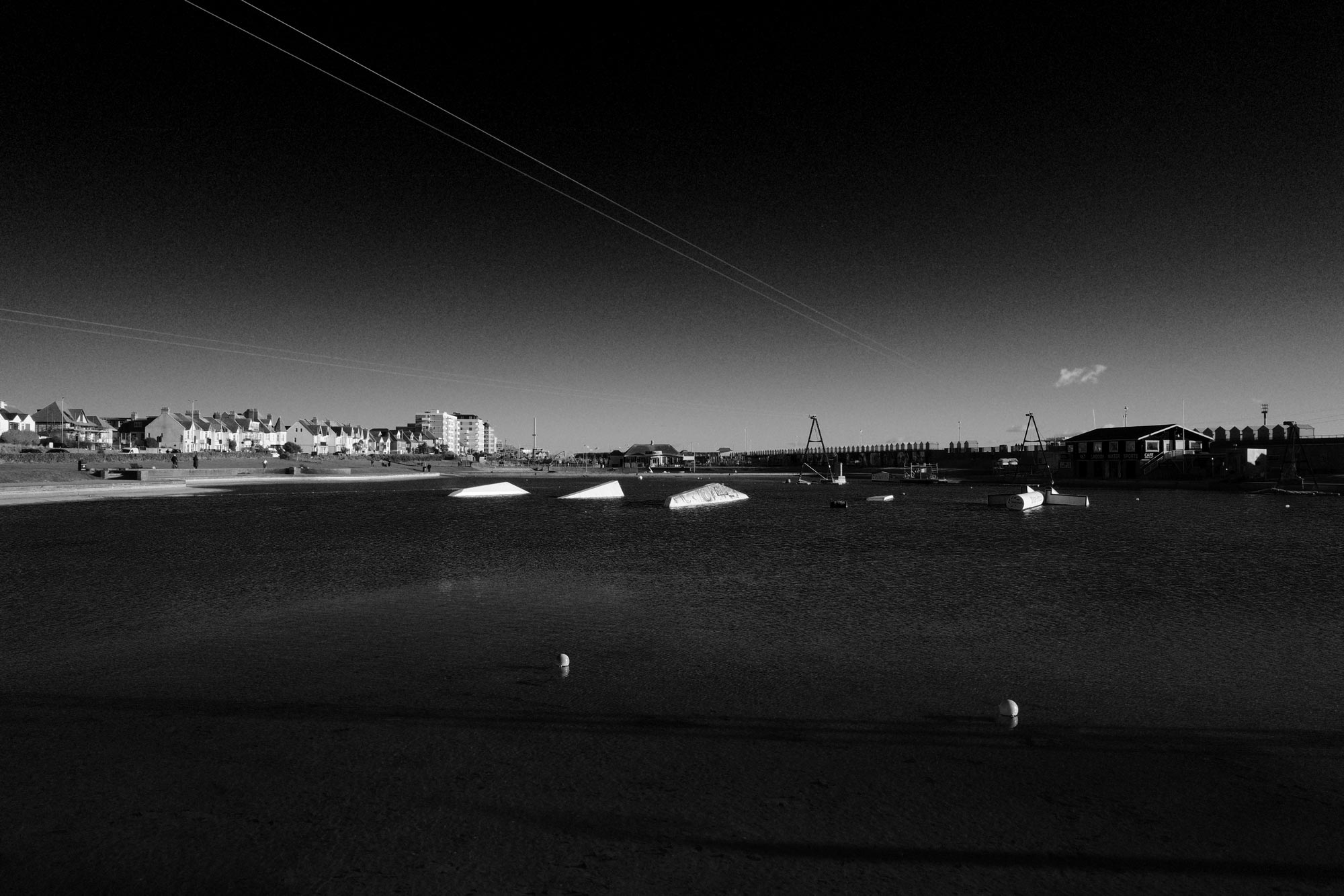 A black and white photo of a watersports centre, with a large stretch of water in the centre, showing houses in the far distance. Using the Hard Monotone film simulation filter.
