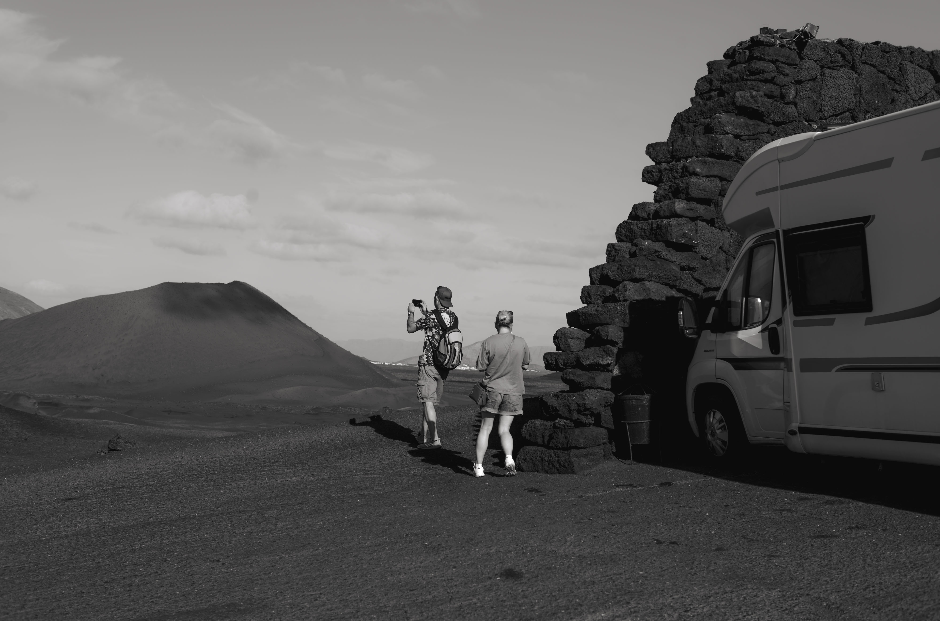 Photo en noir et blanc de deux personnes, debout près d'une caravane et d'un mur, prenant une photo d'une dune de sable, prise avec un Leica M10 Monochrom.