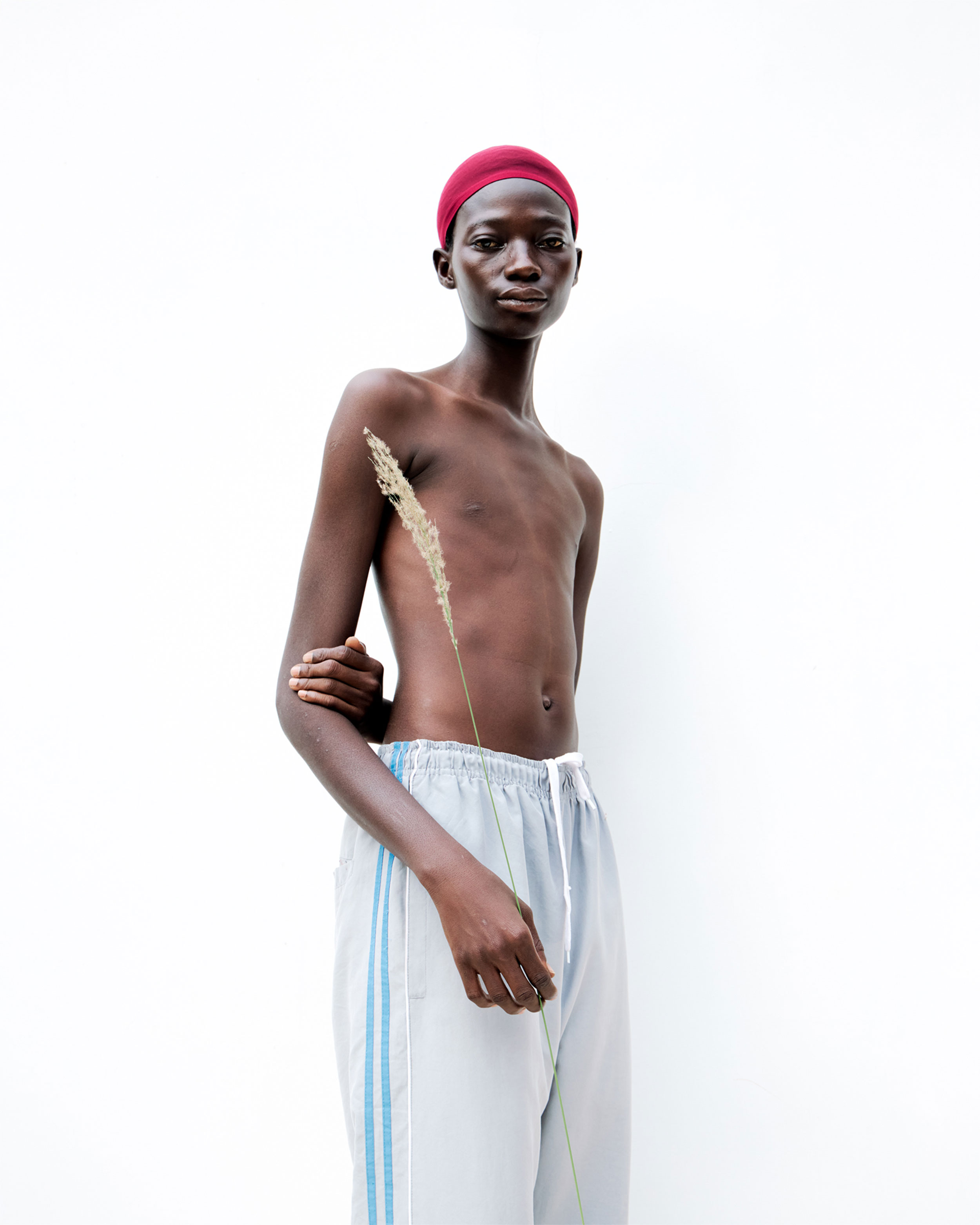 Black person with white sweatpants, red cap, topless, holding an ear of corn in their right hand and looking directly into the camera, shot by Lisa Marie Asubonteng