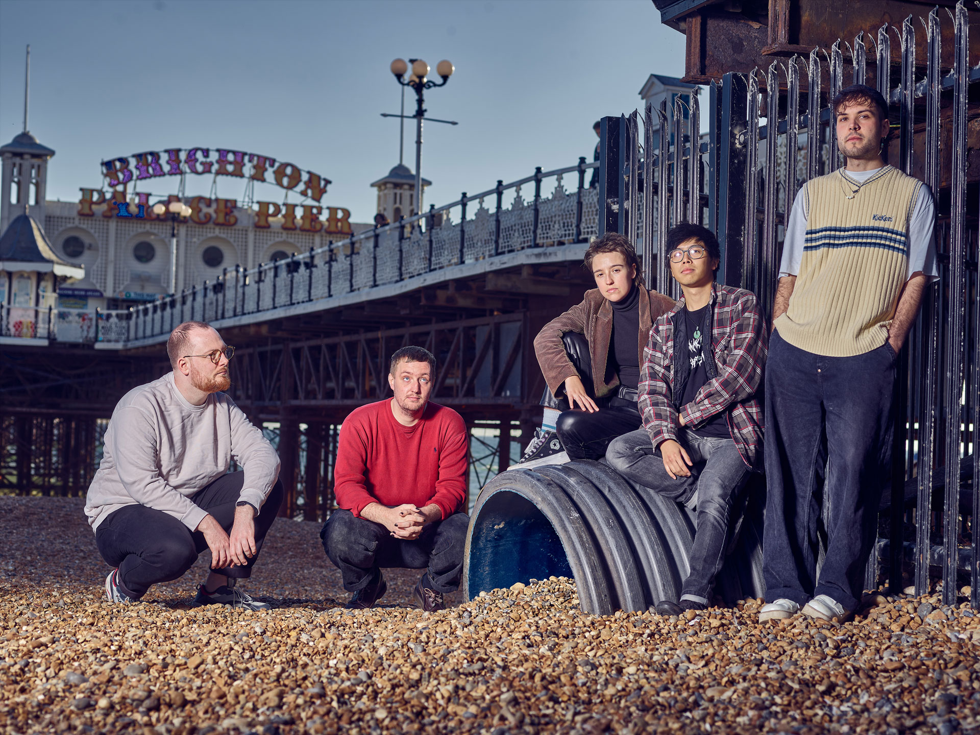 The band ‘Flirting’ sitting on and around a large pipe on Brighton beach, with the Palace Pier, railings and sky in the background, photo by Ki Price