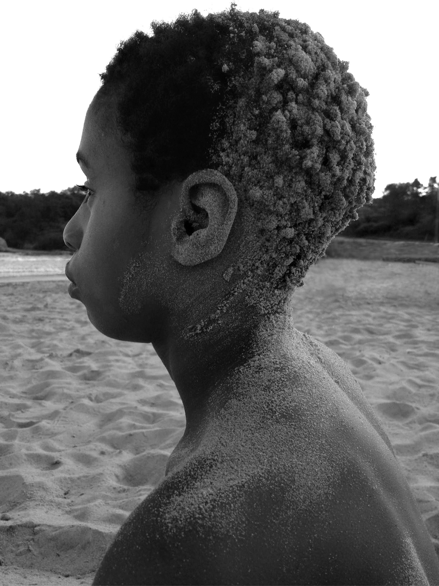 Ein Schwarz-Weiß-Bild eines Jungen, der am Strand sitzt, mit Sand im Nacken, auf den Schultern und am Hinterkopf. Aufgenommen von Kim Fuller.