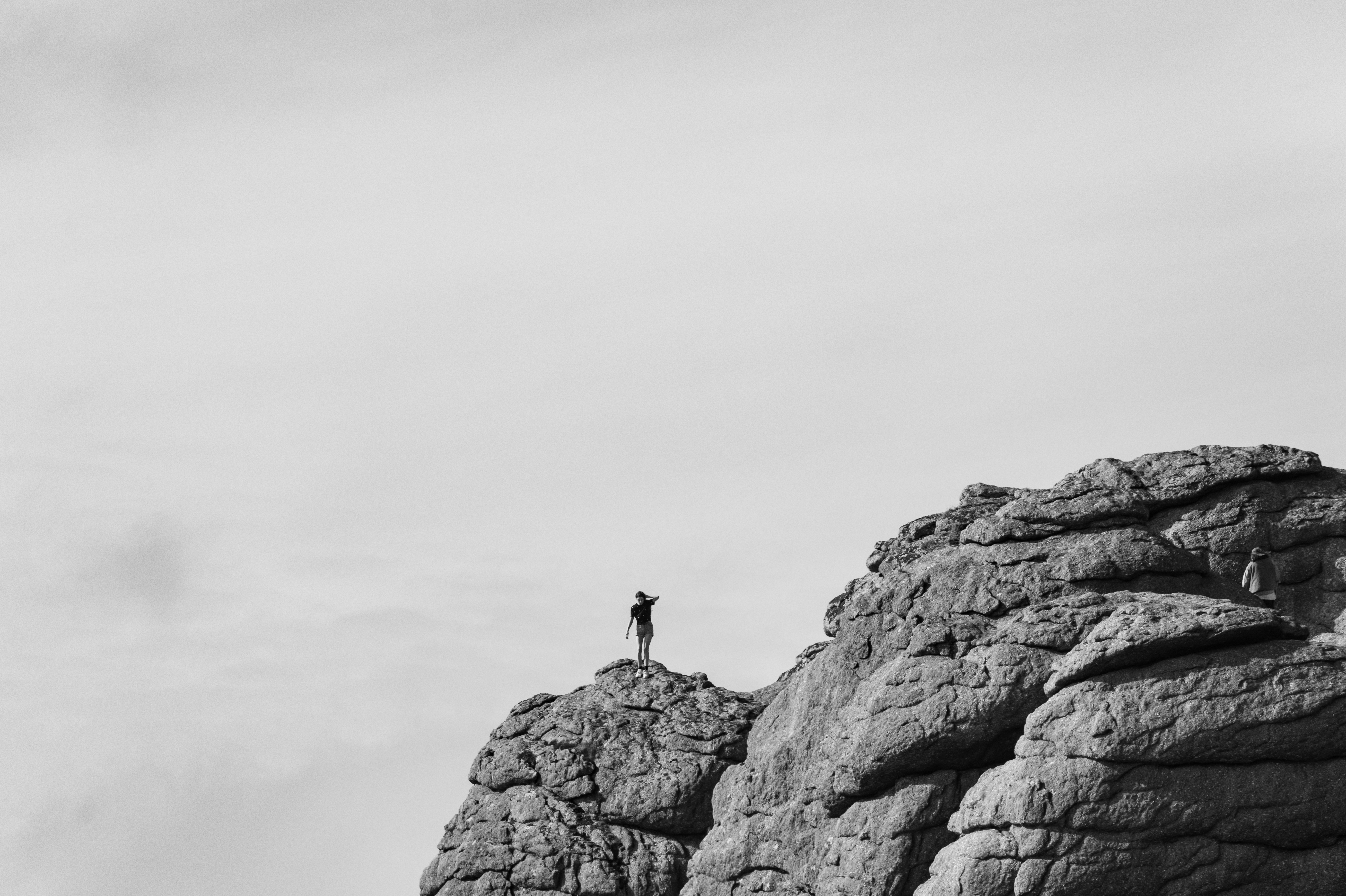 A black-and-white photo of rock formations in Dartmoor National Park. A lone woman stands on top of a rock in the centre of the photo, surrounded by wispy clouds. Photo by Amy Moore with the Panasonic S1R II

