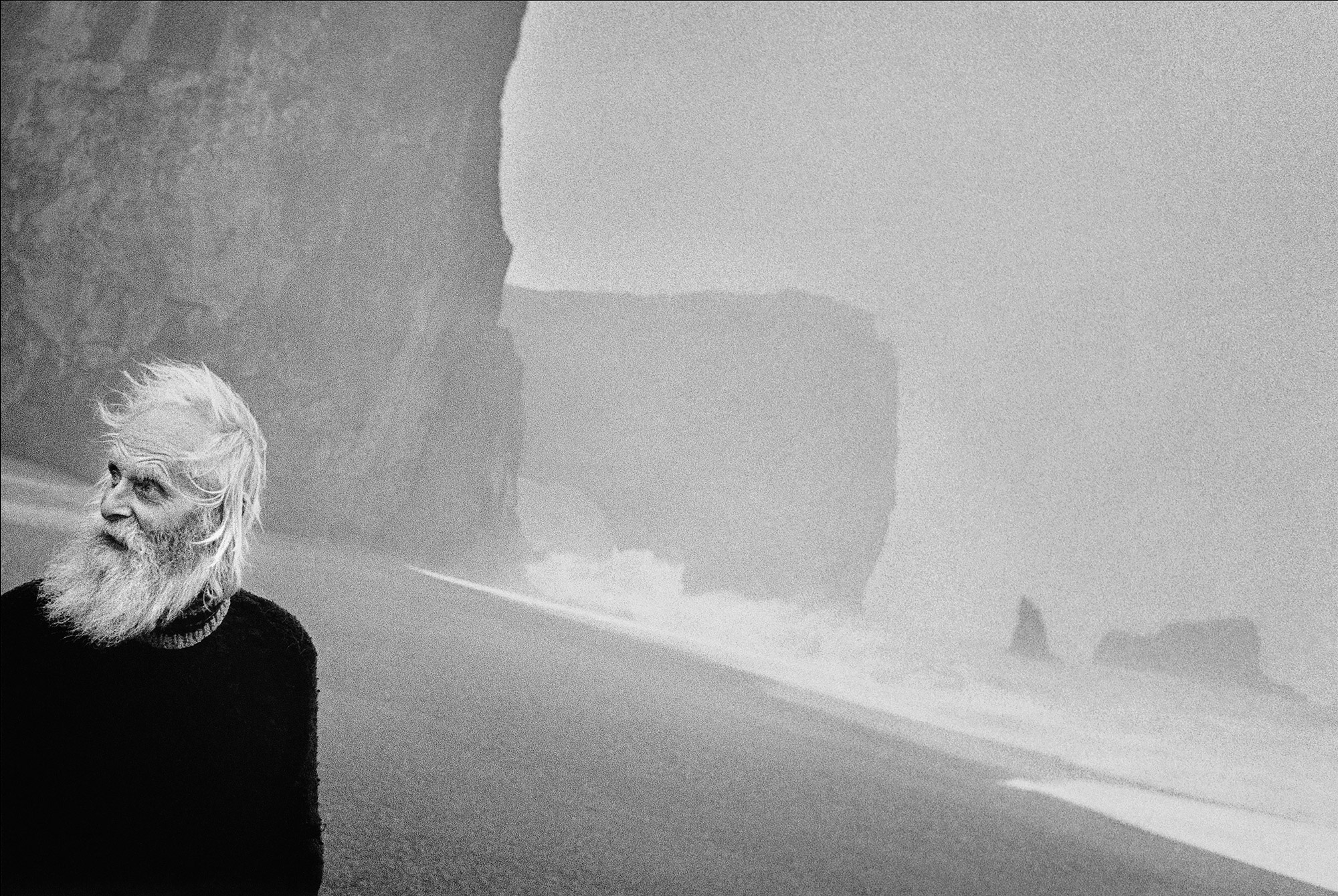Black and white photograph of a person in the front left of the picture looking to the right, mountains and sea in the background, photo taken by Ragnar Axelsson