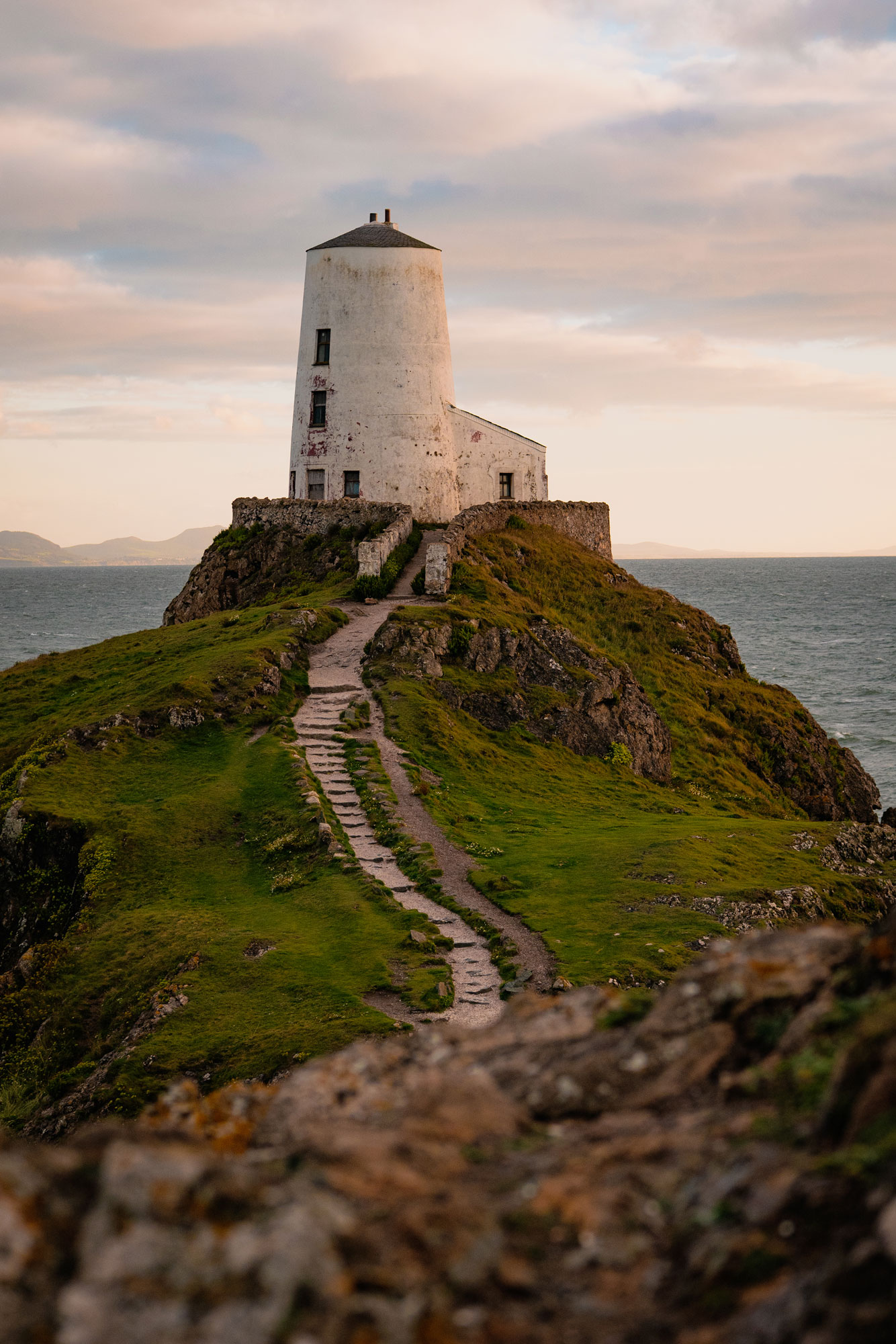 Ein Leuchtturm auf dem höchsten Punkt eines Hügels, zu dem ein Pfad hinaufführt. Das Meer im Hintergrund ist ruhig und blau, der in sanftes Licht getauchte Himmel leicht bewölkt.