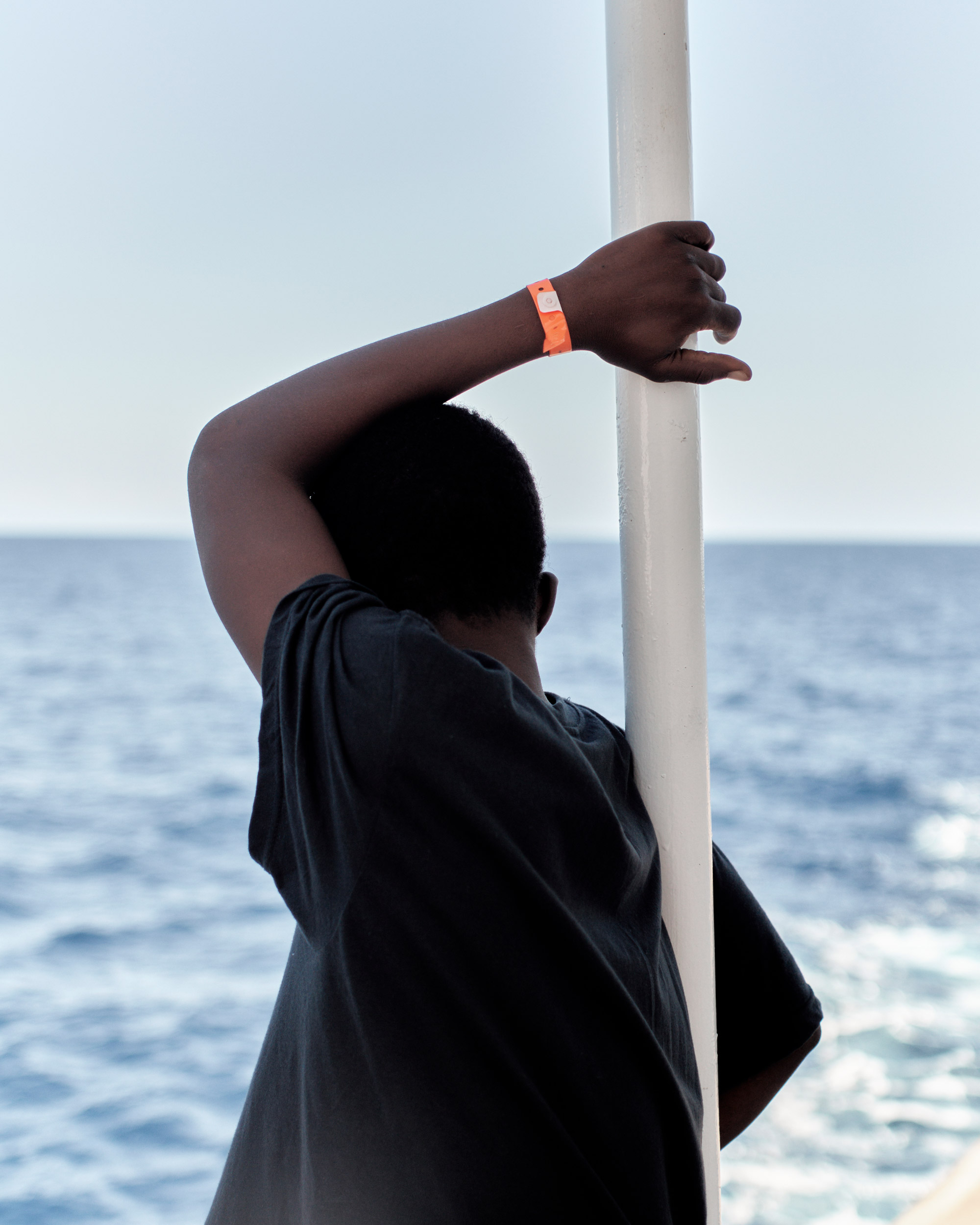 Person, die sich an eine weiße Stange lehnt und sich mit der linken Hand über dem Kopf daran festhält, von hinten fotografiert, am linken Handgelenk ein oranges Armband.