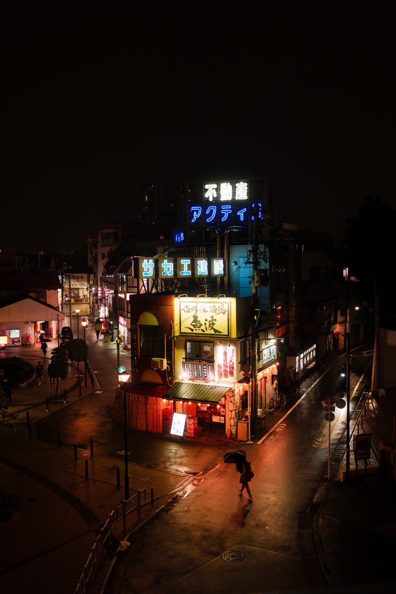 A nighttime view of a Japanese street illuminated by vibrant neon signs and shop lights, with a lone pedestrian walking under an umbrella on a wet street. Photographed by Ruby Cruden with a Sony DSC-RX1 RM II.