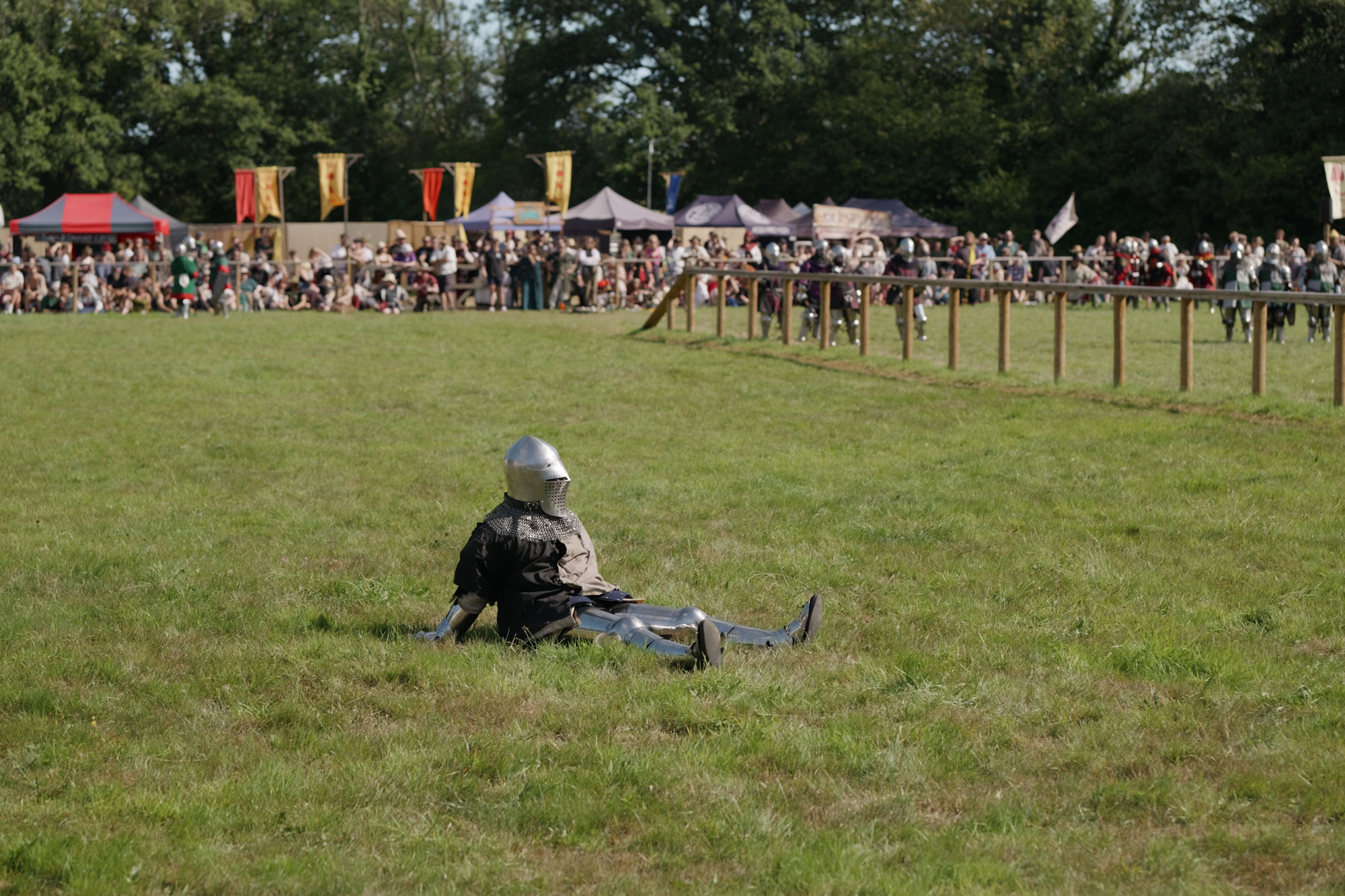 A screenshot from an Open Gate video recording. A person in a knight's costume can be seen on the floor, looking tired after a battle. There is a crowd in the background as well as more people in costume, food tents and decorative medieval-style signs.