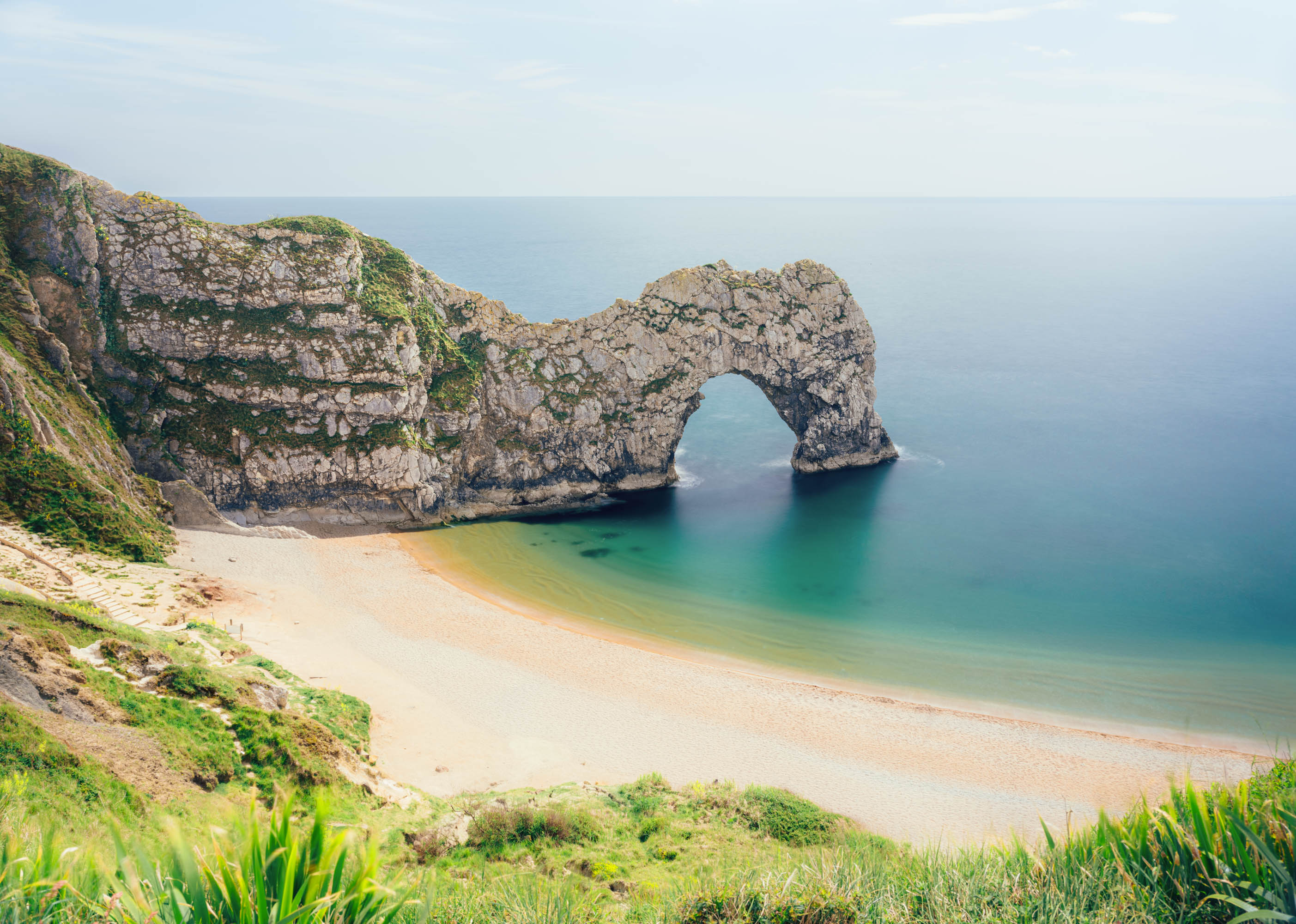 Durdle Door in Dorset, England in the middle of the day, Coastal cliff and natural stone arch over turquoise sea, with a sweeping sandy beach below and grassy foreground on a clear day