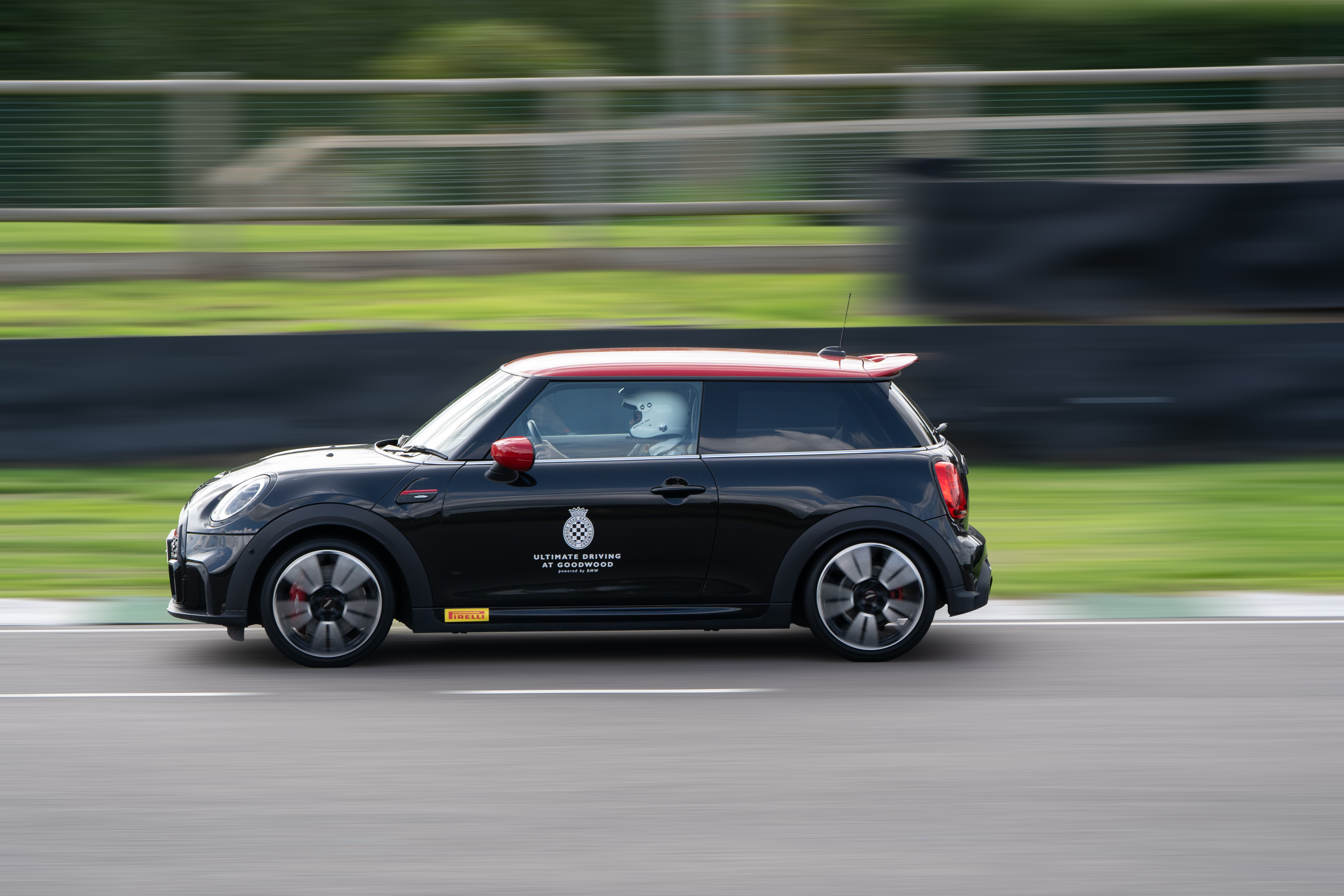 A black Mini Cooper races by, the car in sharp detail and the background out of focus. The driver can be seen through the window wearing a white helmet.