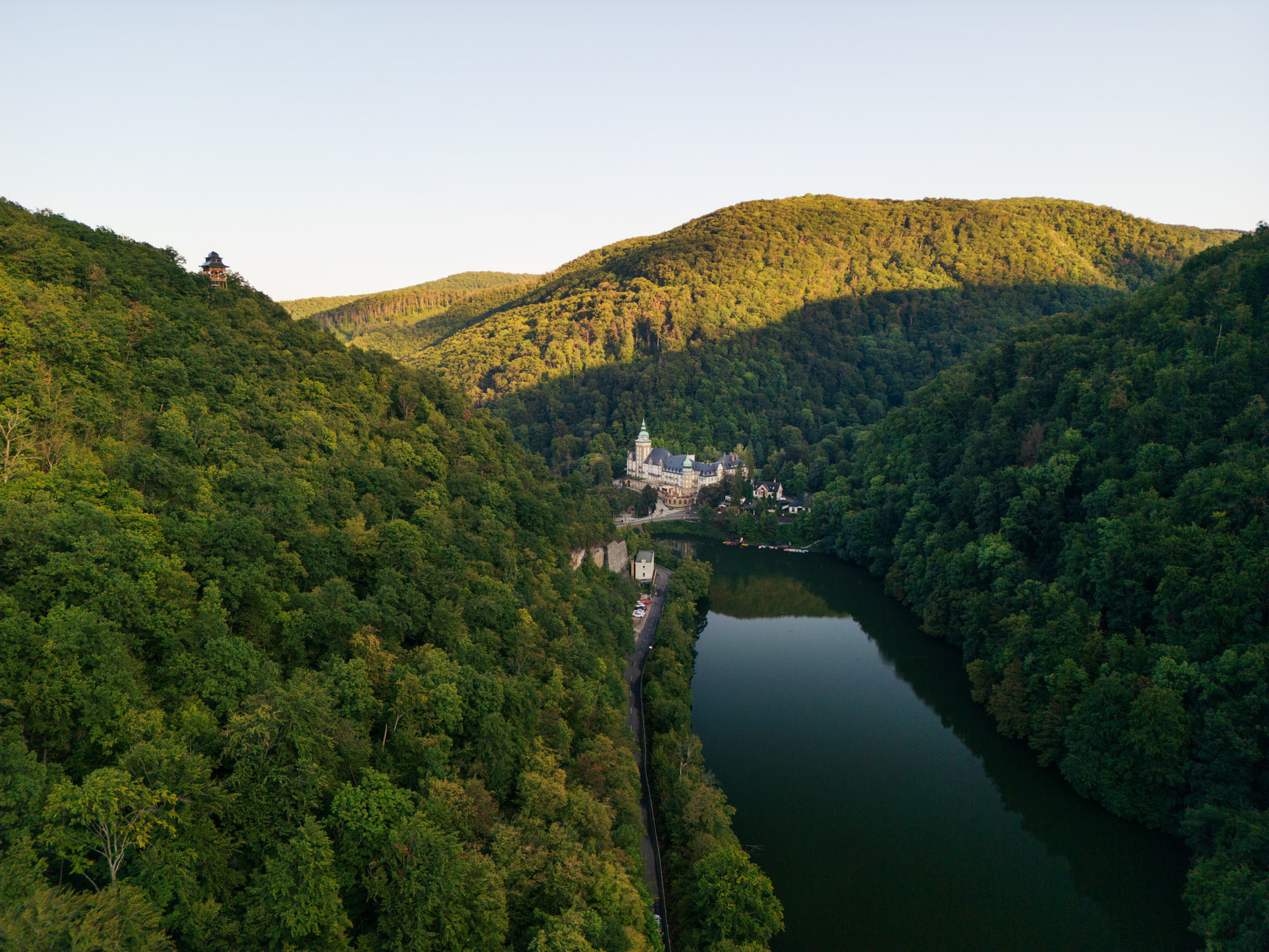 Drohnenaufnahme eines bewaldeten Tals mit einem langgestreckten See und einem historischen Gebäude am Ufer im warmen Abendlicht. Aufgenommen von Adam Makkai mit einer DJI Mini 3 Pro.