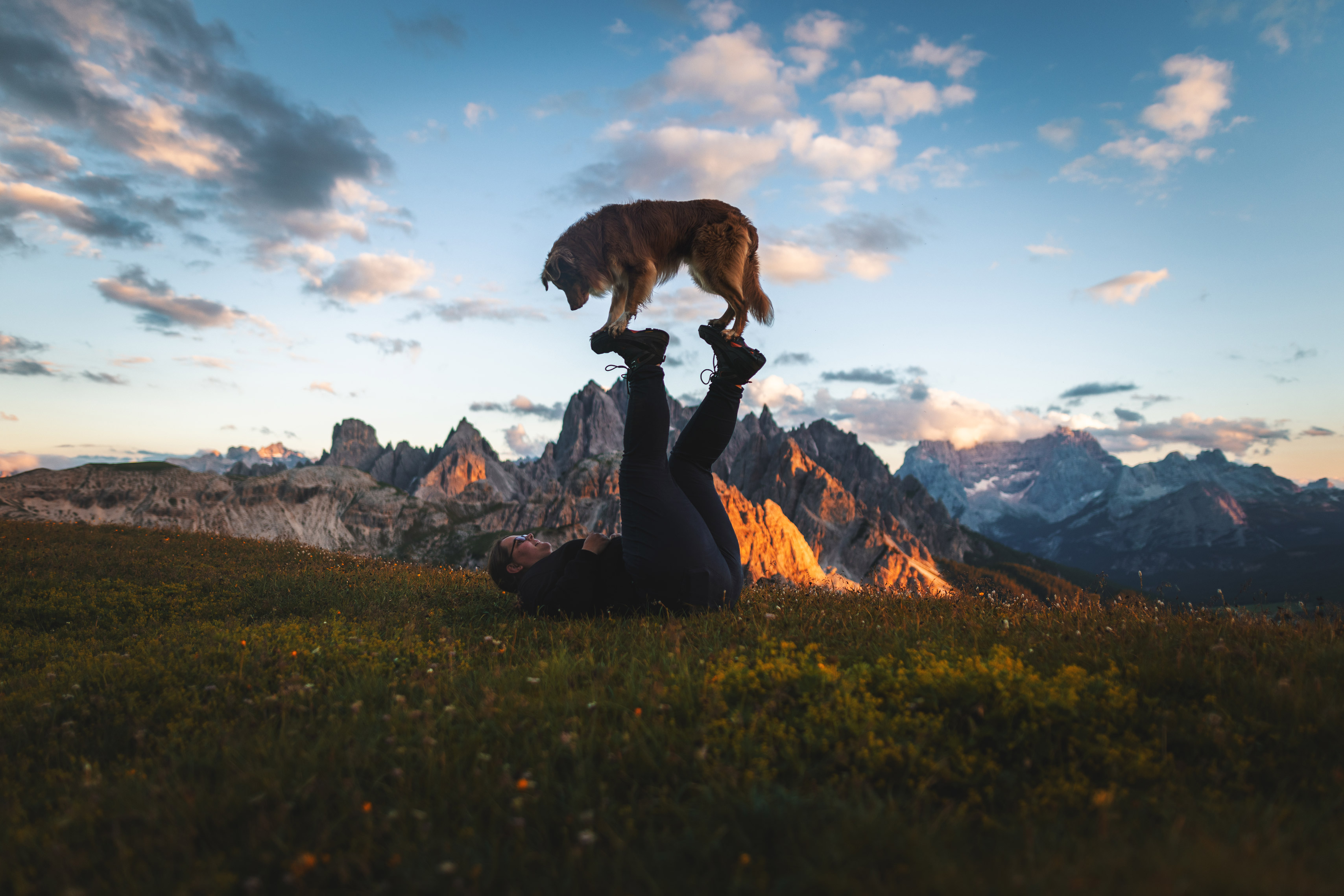 Image of a person lying on the ground with his feet stretched upwards on which a dog is balancing, against a mountain backdrop, by Lea Heidt