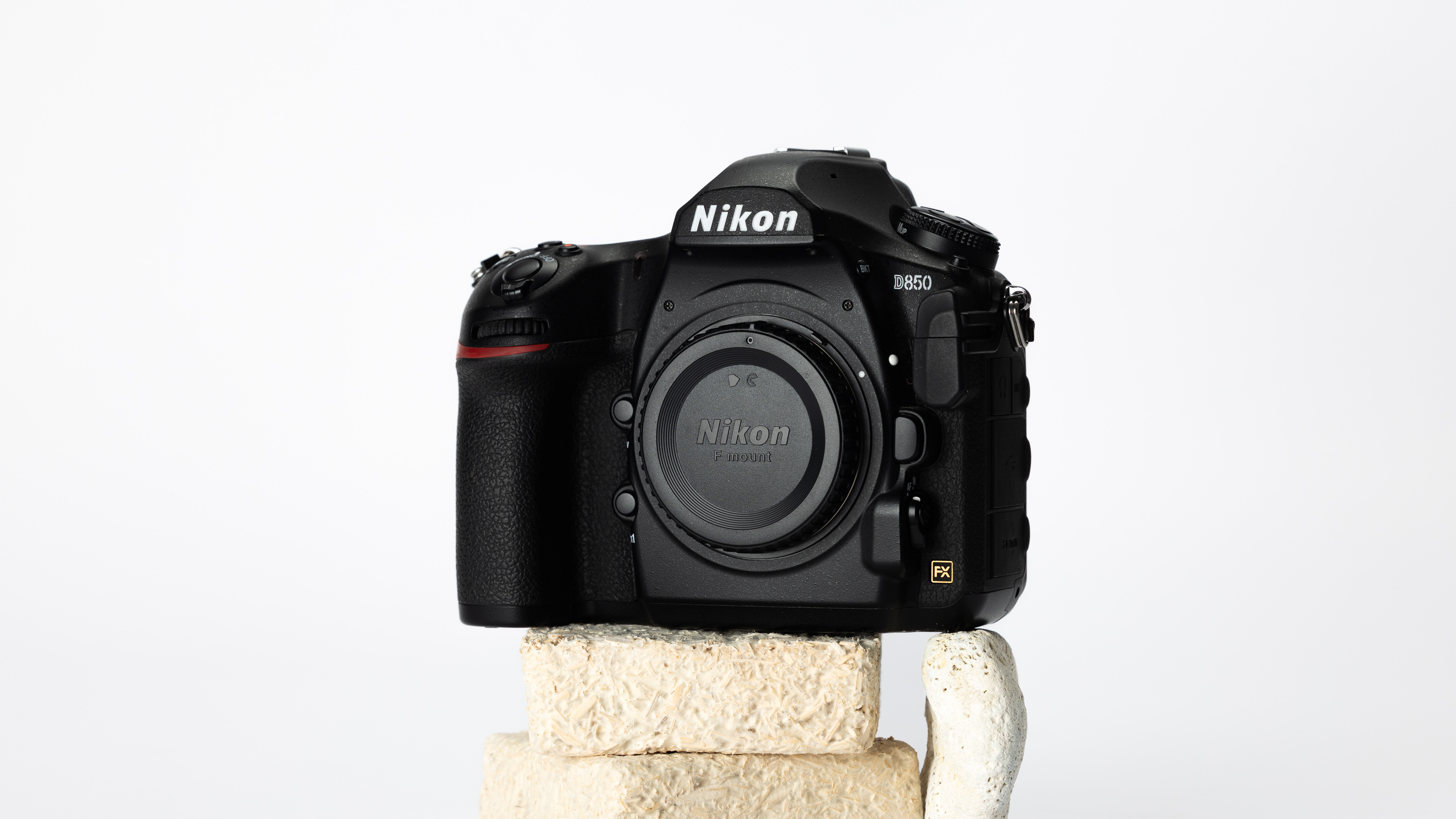 A product shot of a secondhand Nikon D850 camera propped up on a rocky podium on an off white background.