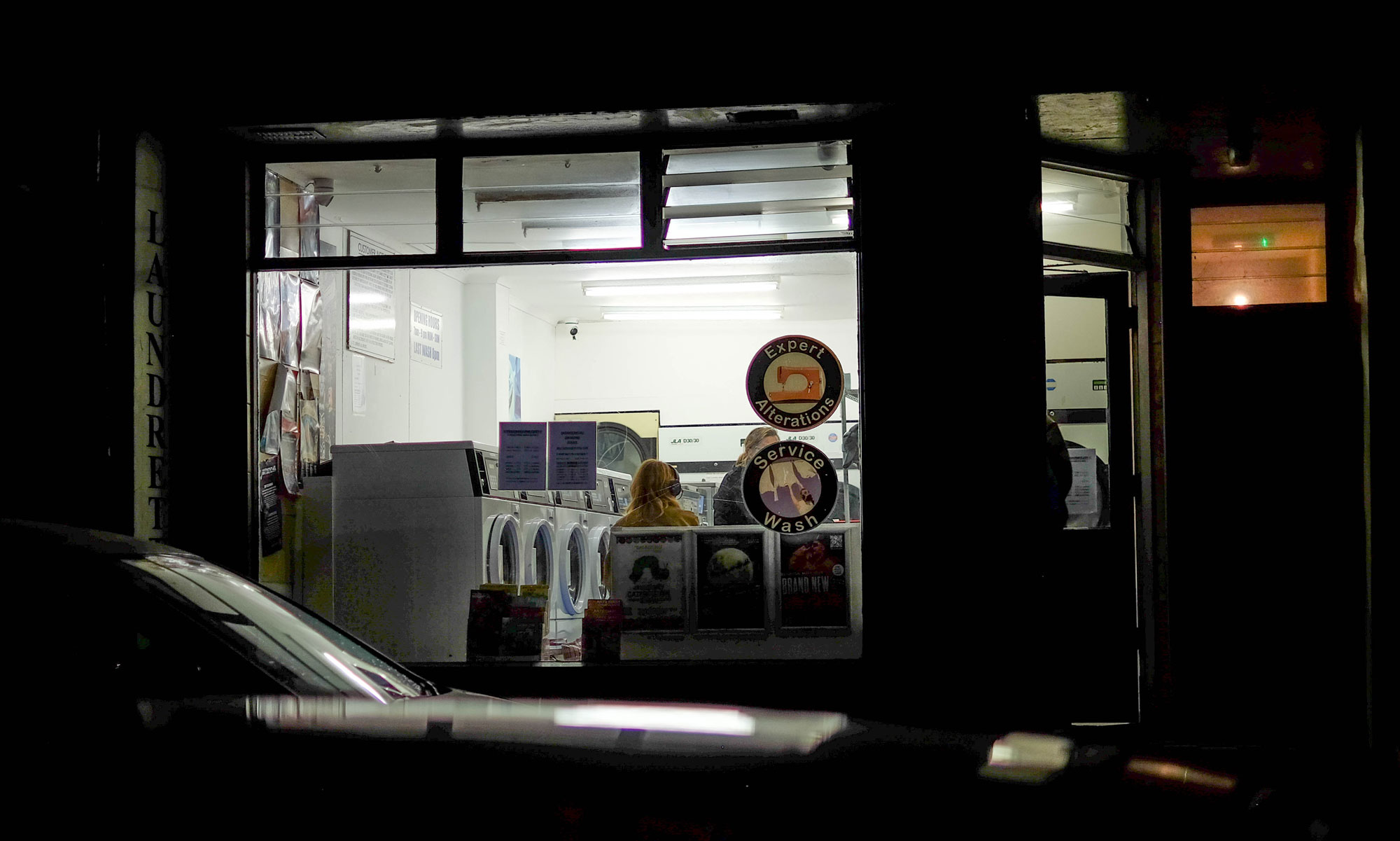 In the evening, a person wearing headphones waits inside a laundrette, visible through the window and lit by bright fluorescent lights.
