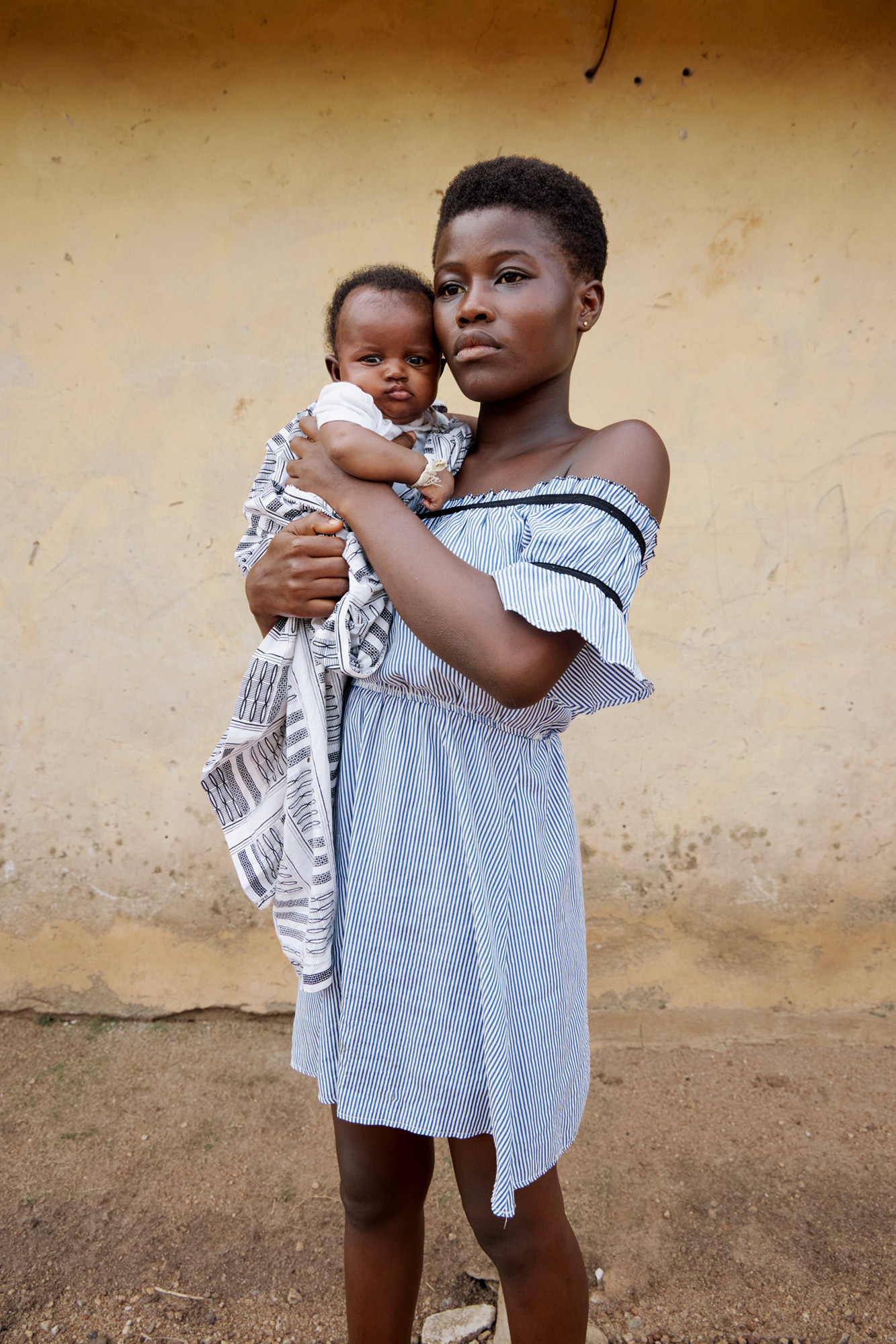 A parent wearing a white dress holding their baby in front of a wall, shot by Lisa Marie Asubonteng with a Canon EOS R