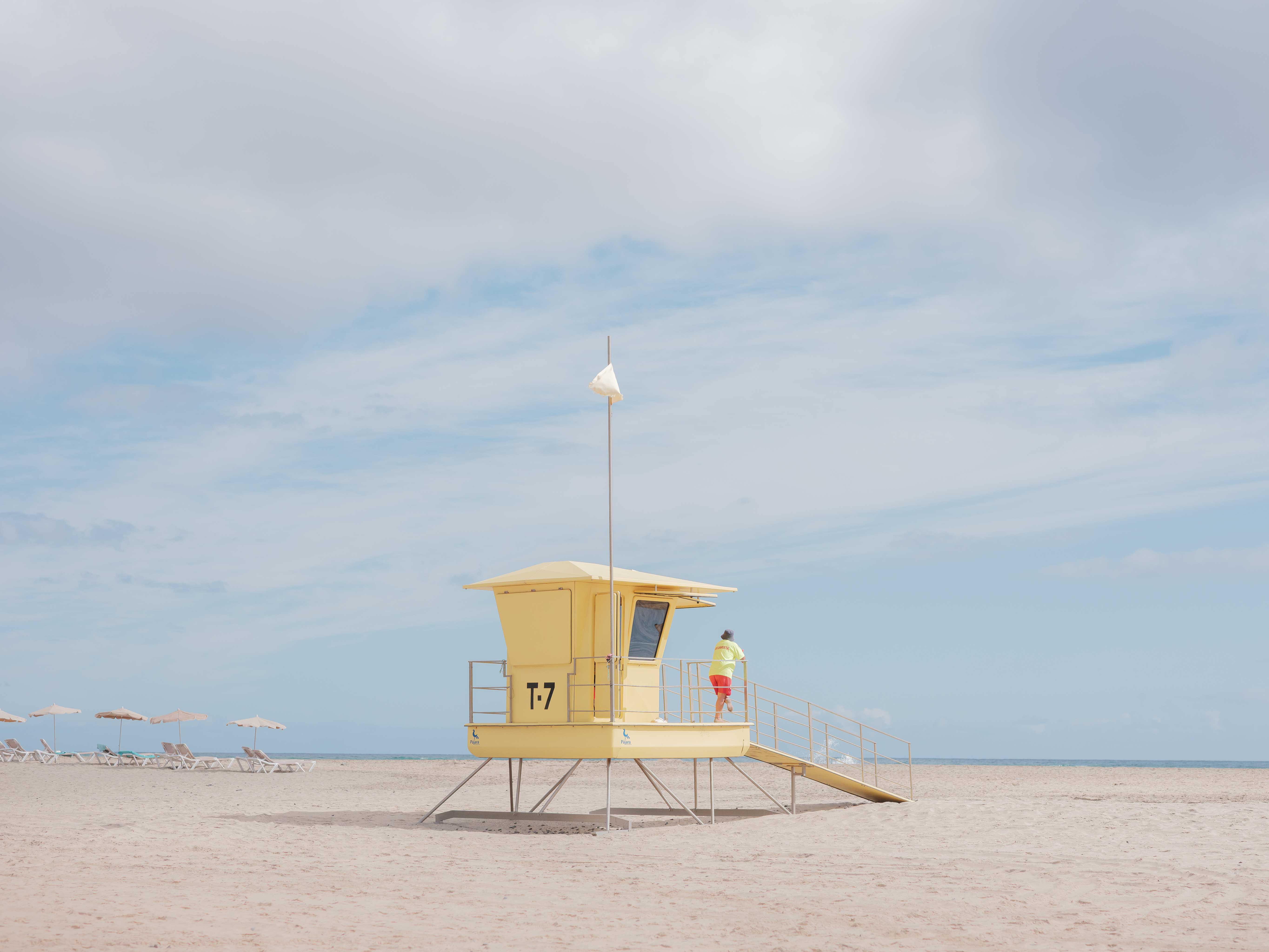 A yellow lifeguard hut on the beach with a lifeguard looking out to sea. By Connor Redmond.