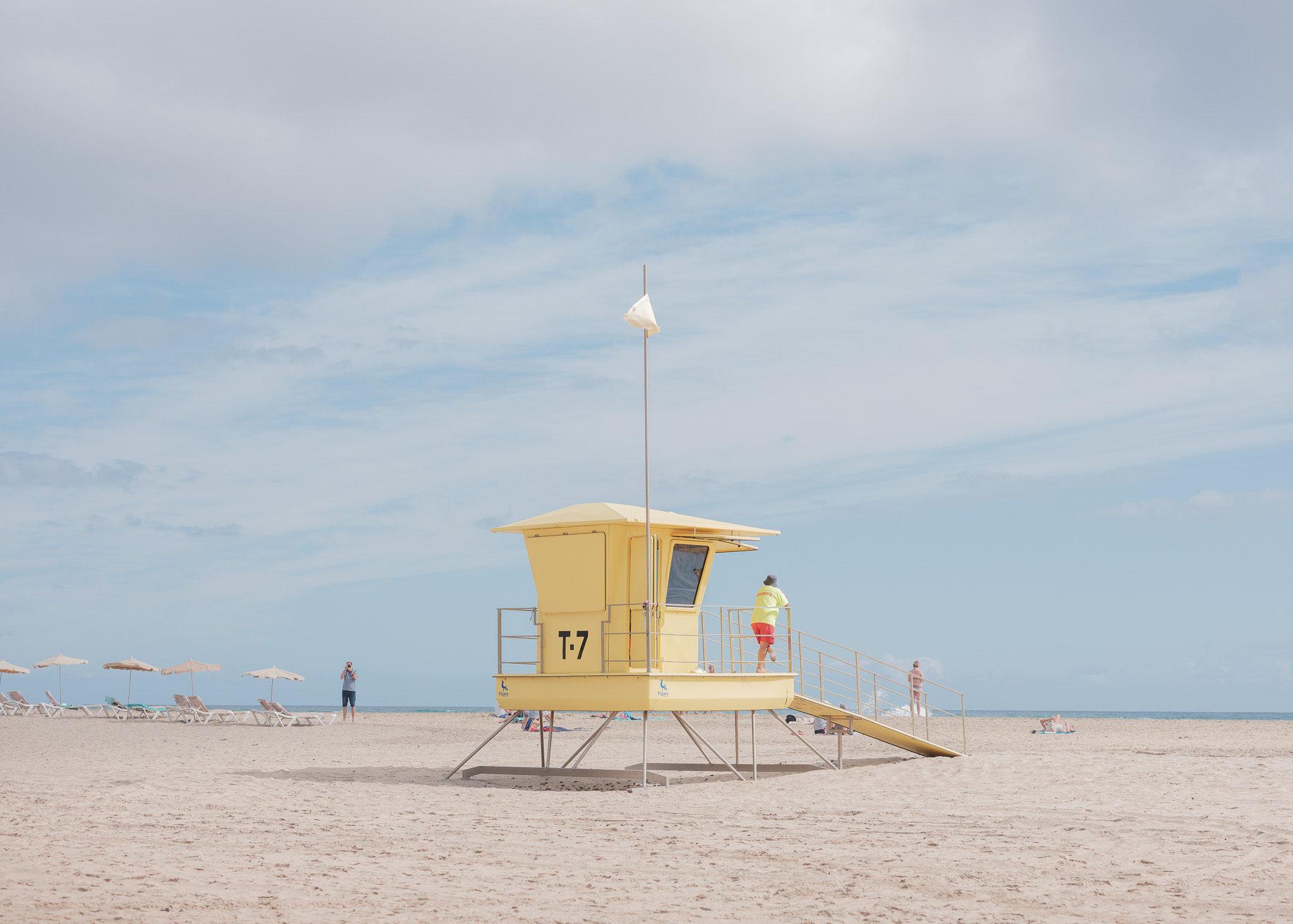 Eine gelbe Rettungsschwimmerhütte am Strand mit einem Rettungsschwimmer, der auf das Meer hinausschaut. Von Connor Redmond.

