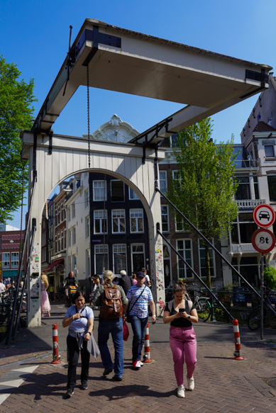 Een levendig straatbeeld op de Amsterdamse Armbrug, waar locals en bezoekers de iconische brug oversteken in helder voorjaarslicht, gefotografeerd door Louise Kluit
