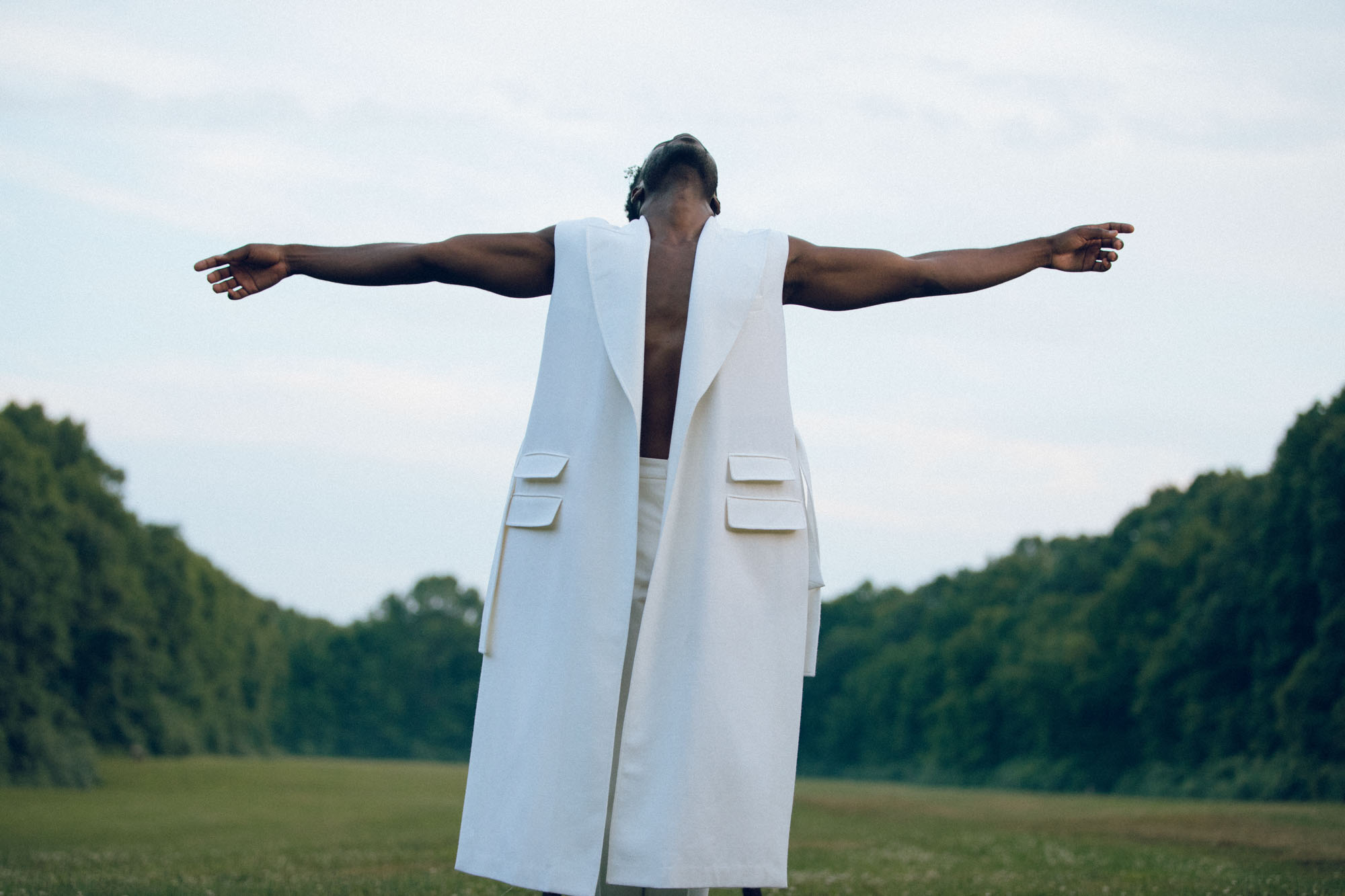 Portrait of a model with their arms outstretched, wearing a white sleeveless jacket, surrounded by trees, conveying a sense of freedom 