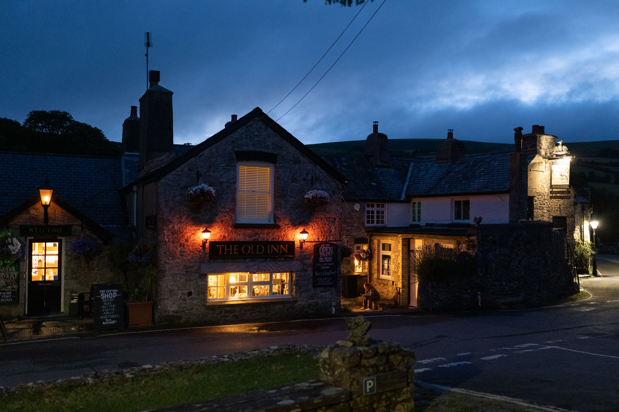 A pub with a sign that reads ‘The Old Inn’ at dusk. The pub lights are lighting the road in front of it with a warm glow.
