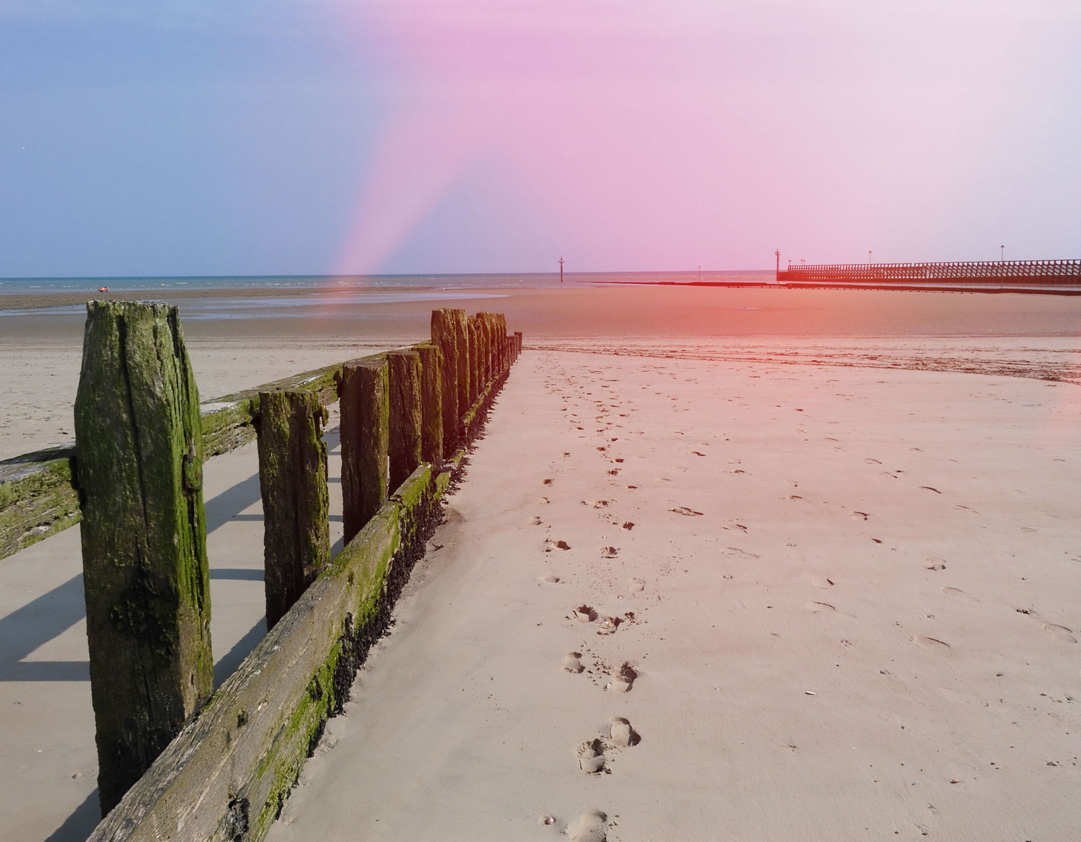 The beach at low tide, with footprints in the sand by the wooden groynes. The photo has a light leak effect coming from the top right corner. 
