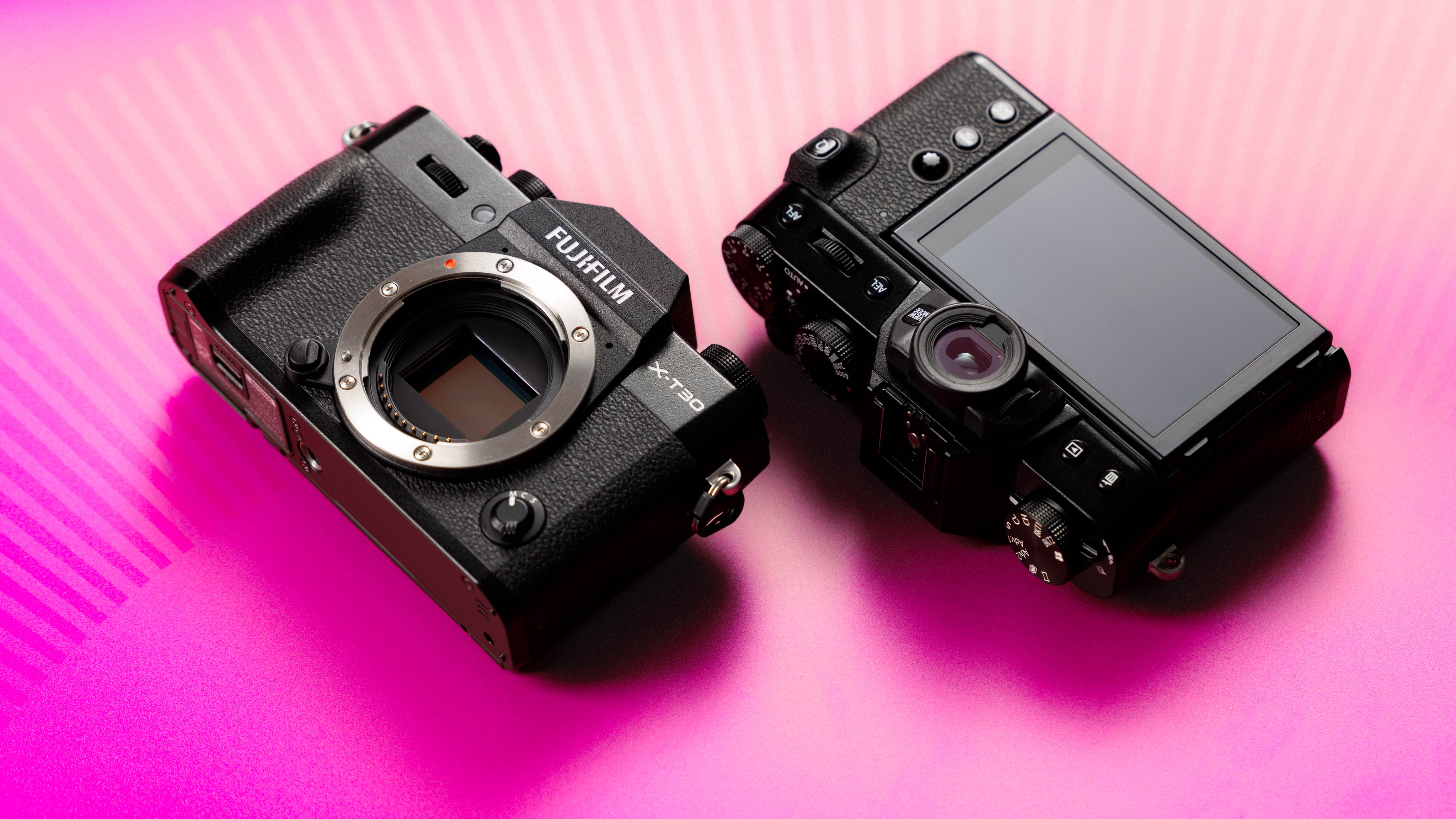 Fujifilm X-T30 mirrorless camera body shown from front and rear angles on a vibrant pink background, highlighting sensor mount, screen and control layout.