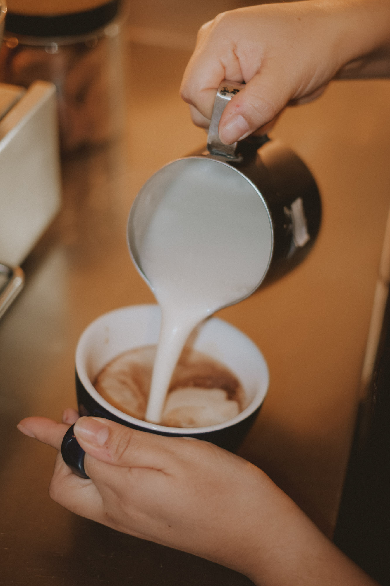 Close-up of hands pouring steamed milk into a coffee cup, creating latte art with smooth foam in a warm café setting.