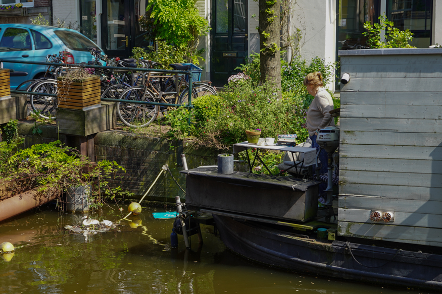 Een rustig moment op een Amsterdams woonbootterras, waar een vrouw uitkijkt over de gracht, omringd door planten en fietsen, gefotografeerd door Louise Kluit