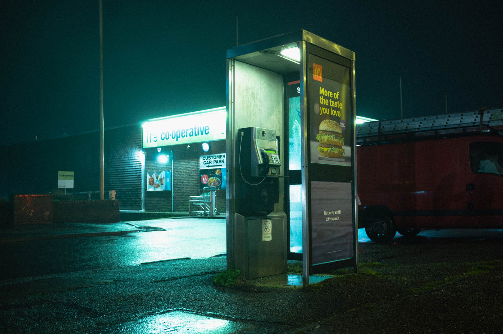 Eine Telefonzelle bei Nacht mit dem hellen Licht eines Lebensmittelgeschäfts im Hintergrund. Aufgenommen von Jakub Golis.