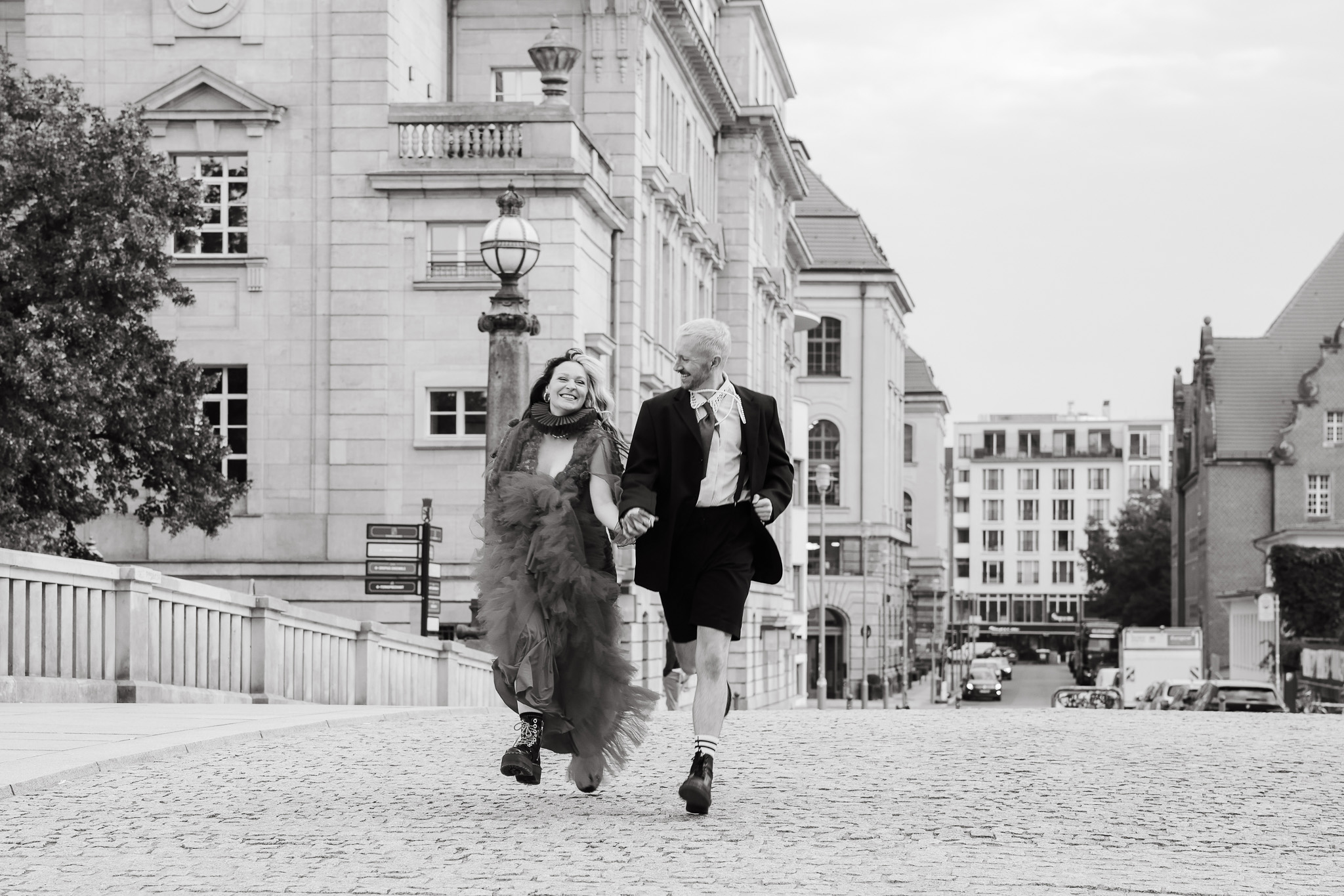 A black-and-white wedding scene of a boldly dressed couple walking cheerfully along a cobbled Berlin street. Shot by Marina Polovinkina on a Canon EOS R6 Mark II.