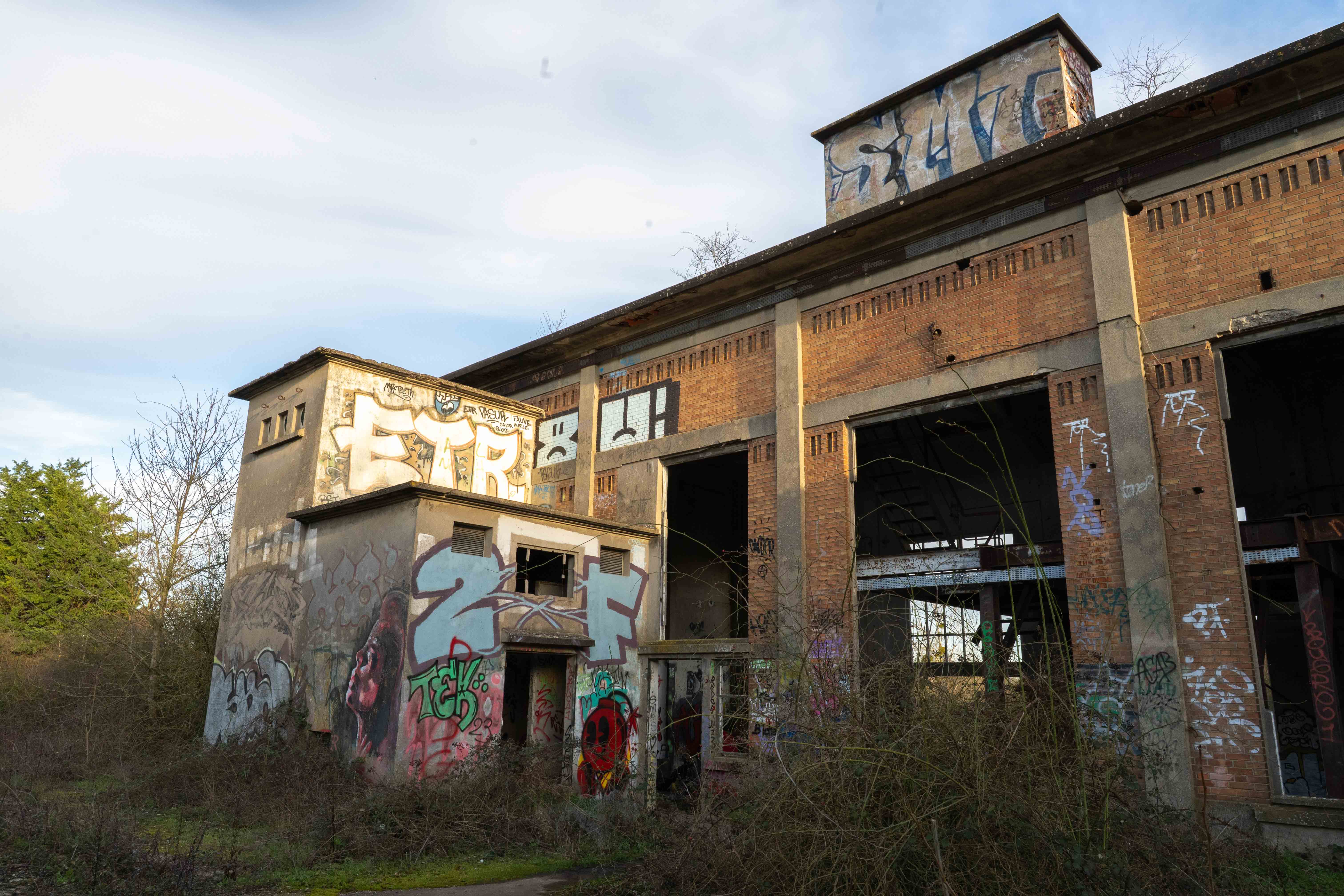 The exterior of a large abandoned brick building covered in colourful graffiti, surrounded by overgrown vegetation beneath a bright sky. Photo taken by Lukas Davos.