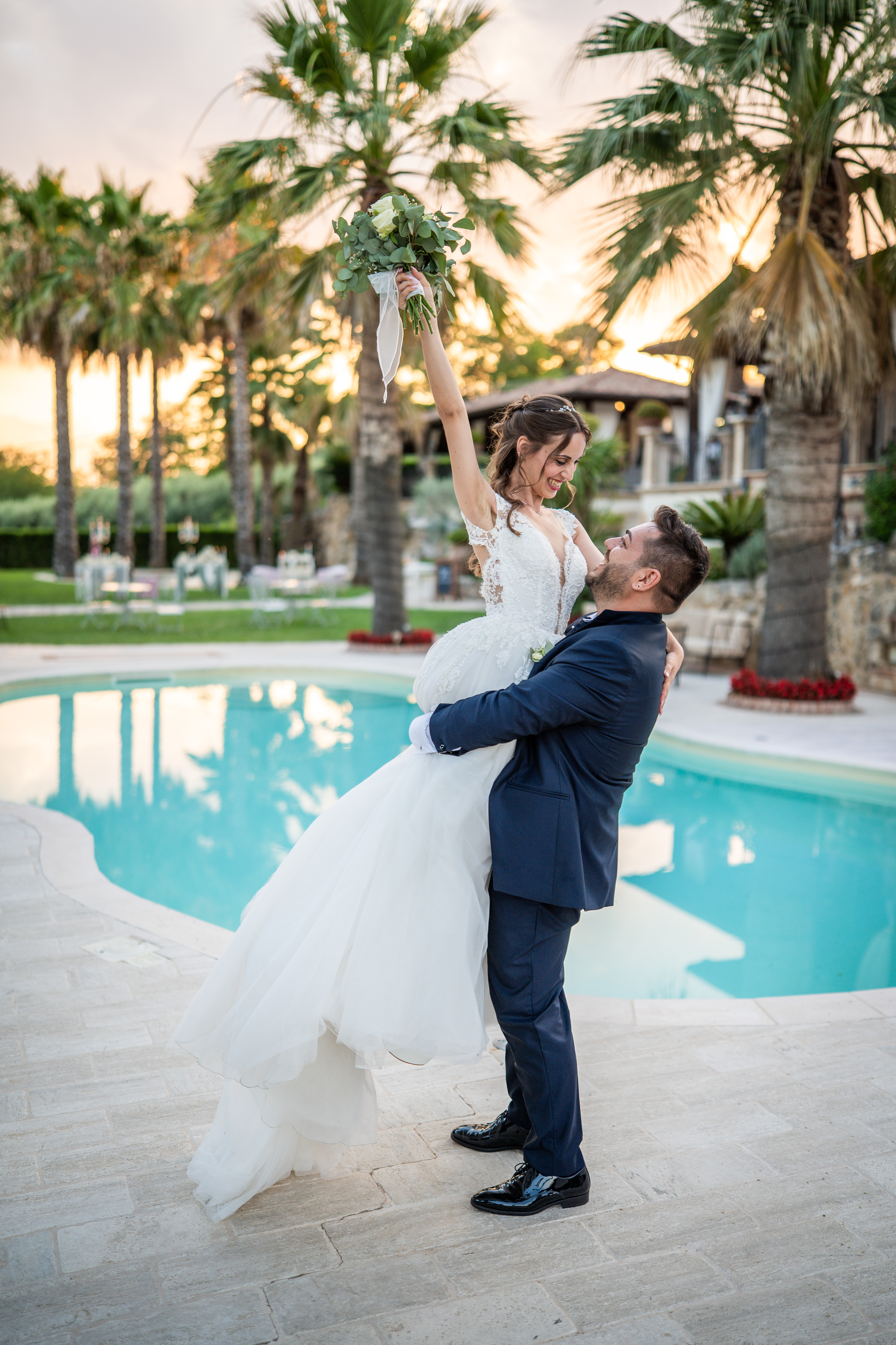 Sposo che solleva la sposa in braccio a bordo piscina, lei ride con il bouquet alzato, tramonto e palme sullo sfondo.