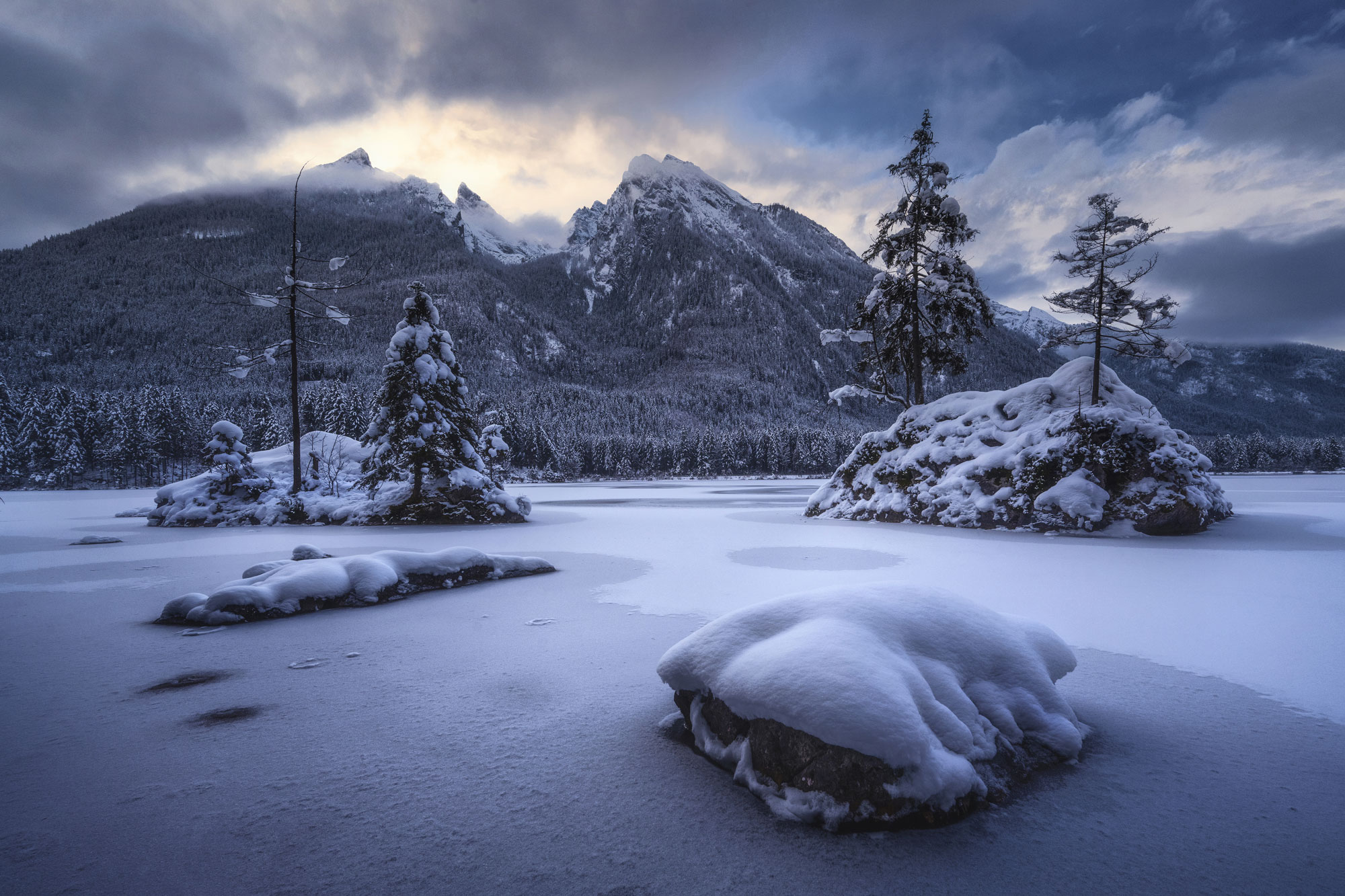 Schneelandschaft mit kleinen Hügeln und Tannen im Vordergrund und hohen Bergen im Hintergrund, hinter denen die Sonne die Wolken durchbricht, aufgenommen von Kilian Schönberger

